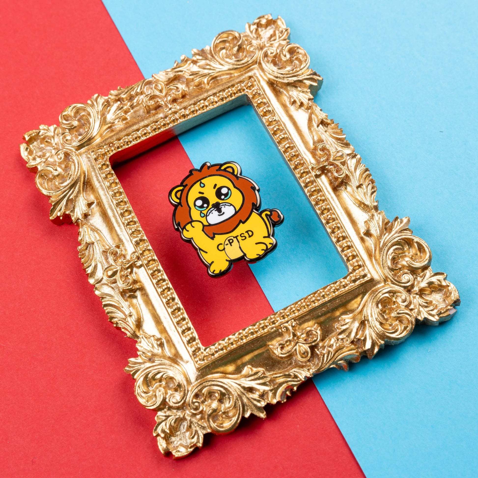 The Complex Post Troarmatic Stress Disorder Lion Enamel Pin - C-PTSD - Complex Post Traumatic Stress Disorder on red and blue card inside a gold ornate frame. A yellow crying smiling and sweating male lion with its paw raised in the middle with 'C-PTSD' across its chest. The enamel pin design is raising awareness for complex post traumatic stress disorder.