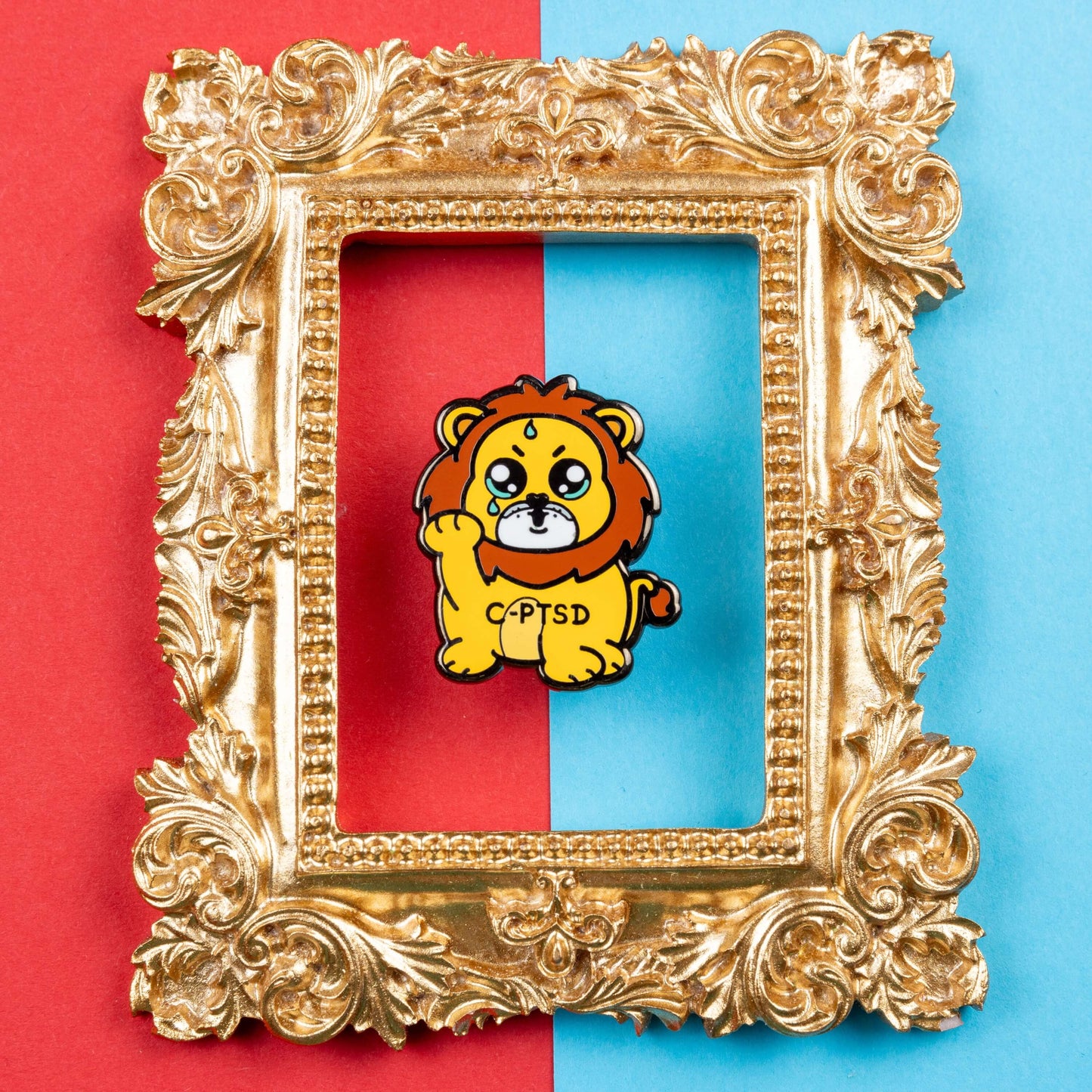 The Complex Post Troarmatic Stress Disorder Lion Enamel Pin - C-PTSD - Complex Post Traumatic Stress Disorder on red and blue card inside a gold ornate frame. A yellow crying smiling and sweating male lion with its paw raised in the middle with 'C-PTSD' across its chest. The enamel pin design is raising awareness for complex post traumatic stress disorder.