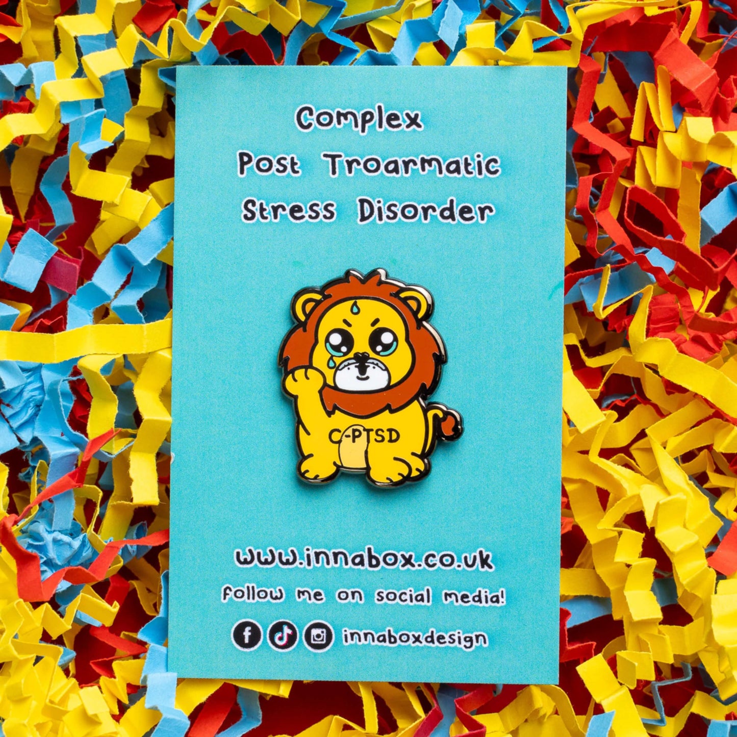 The Complex Post Troarmatic Stress Disorder Lion Enamel Pin - C-PTSD - Complex Post Traumatic Stress Disorder on blue backing card laid on a red, yellow and blue card confetti background. A yellow crying smiling and sweating male lion with its paw raised in the middle with 'C-PTSD' across its chest. The enamel pin design is raising awareness for complex post traumatic stress disorder.