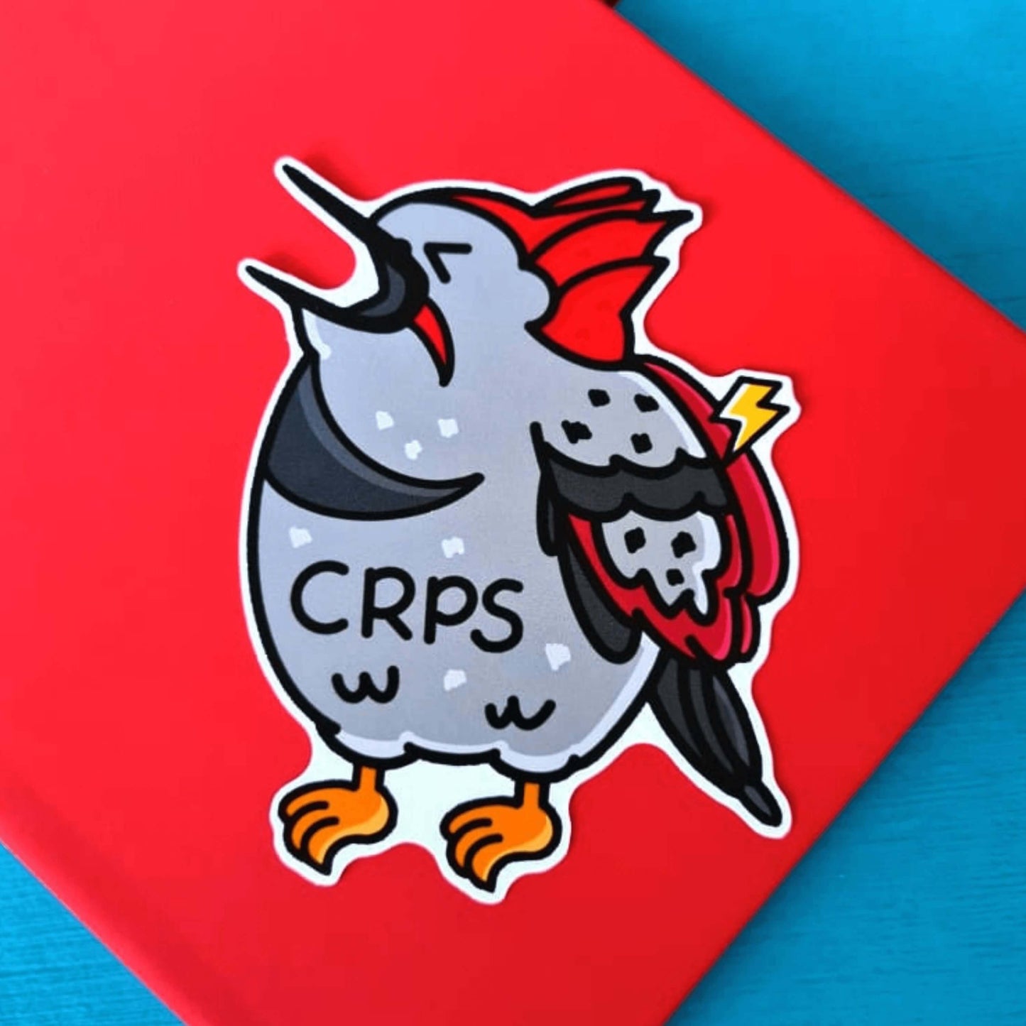 The Compecks Regional Pain Syndrome Sticker - CRPS - Complex Regional Pain Syndrome on a red and blue background. The sticker features a screaming upset woodpecker bird with a lightning bolt on its wing and 'CRPS' written across its belly in black. The design is raising awareness for complex regional pain syndrome.