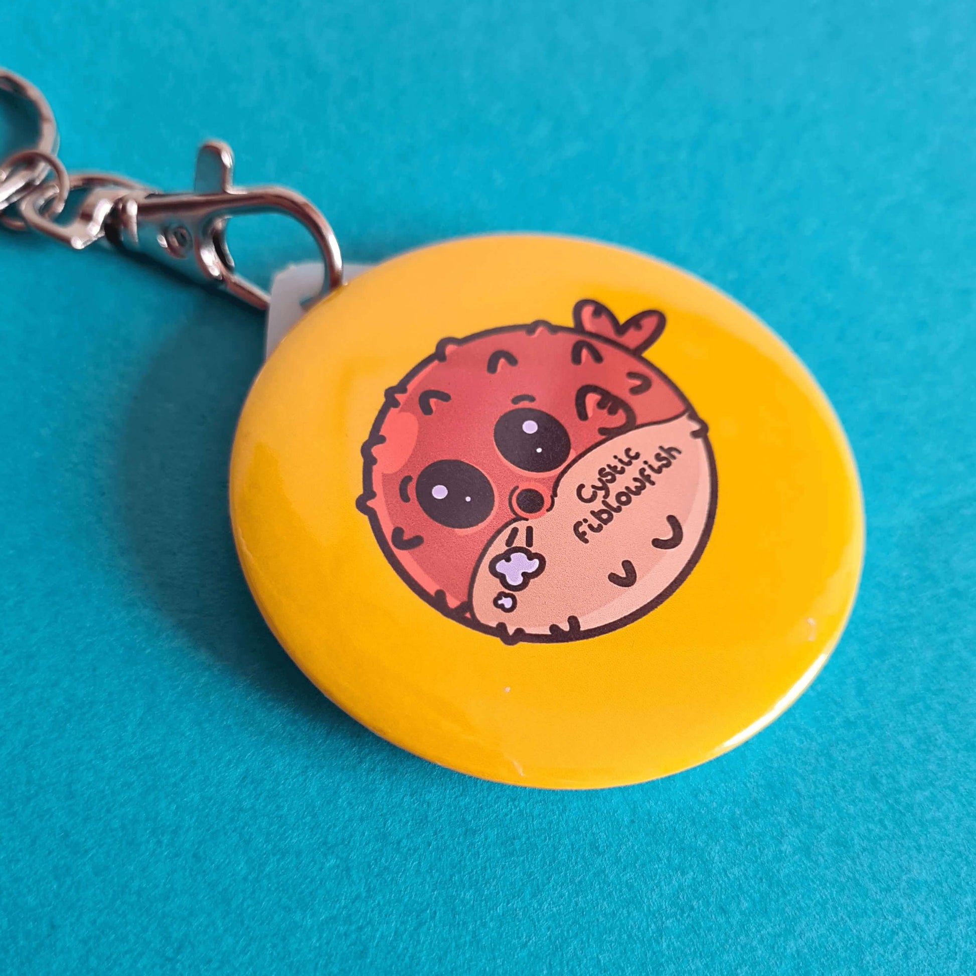 The Cystic Fiblowfish Keyring - Cystic Fibrosis on a blue background. The silver clip plastic yellow circle keychain with a puffer fish blowfish wheezing with 'cystic fiblowfish' written in black on its belly. The design is raising awareness for cystic fibrosis.