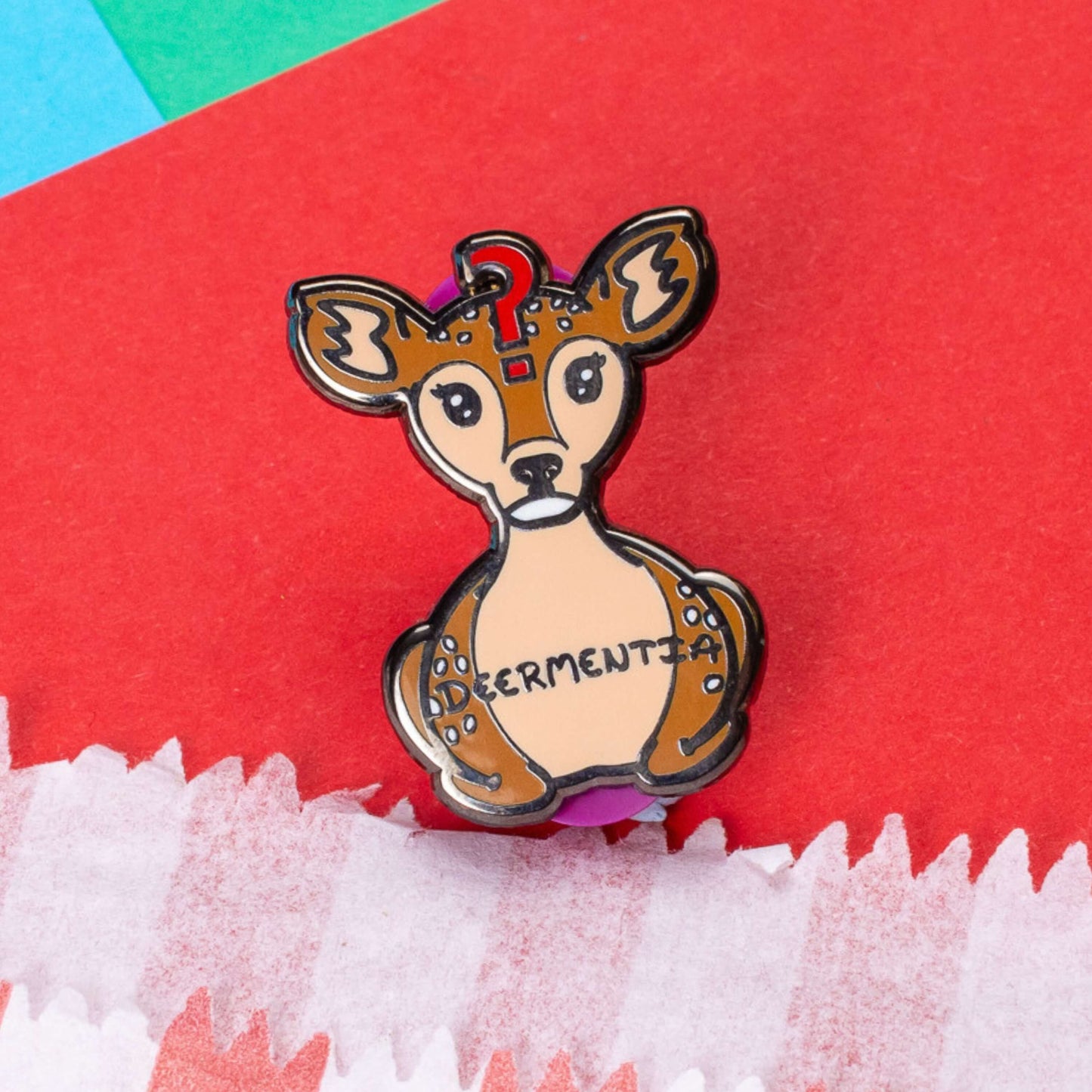 The Deermentia Deer Enamel Pin - Dementia on a red, blue and green card background. A brown female deer laying down with a vacant expression and red question mark above its head with 'deermentia' written across its middle. The hand drawn design is raising awareness for dementia.