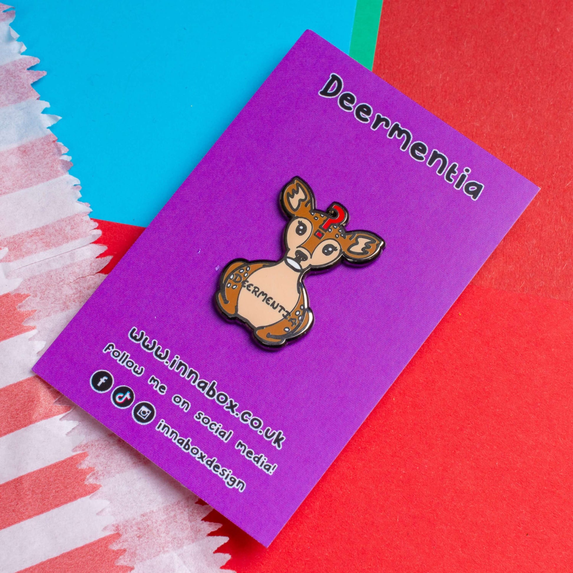 The Deermentia Deer Enamel Pin - Dementia on purple backing card laid on a red, blue and green card background. A brown female deer laying down with a vacant expression and red question mark above its head with 'deermentia' written across its middle. The hand drawn design is raising awareness for dementia.