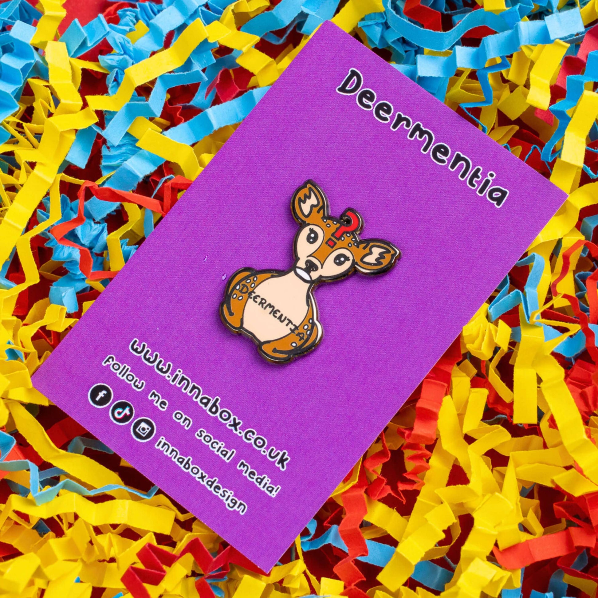 The Deermentia Deer Enamel Pin - Dementia on purple backing card laid on a red, blue and yellow card confetti background. A brown female deer laying down with a vacant expression and red question mark above its head with 'deermentia' written across its middle. The hand drawn design is raising awareness for dementia.