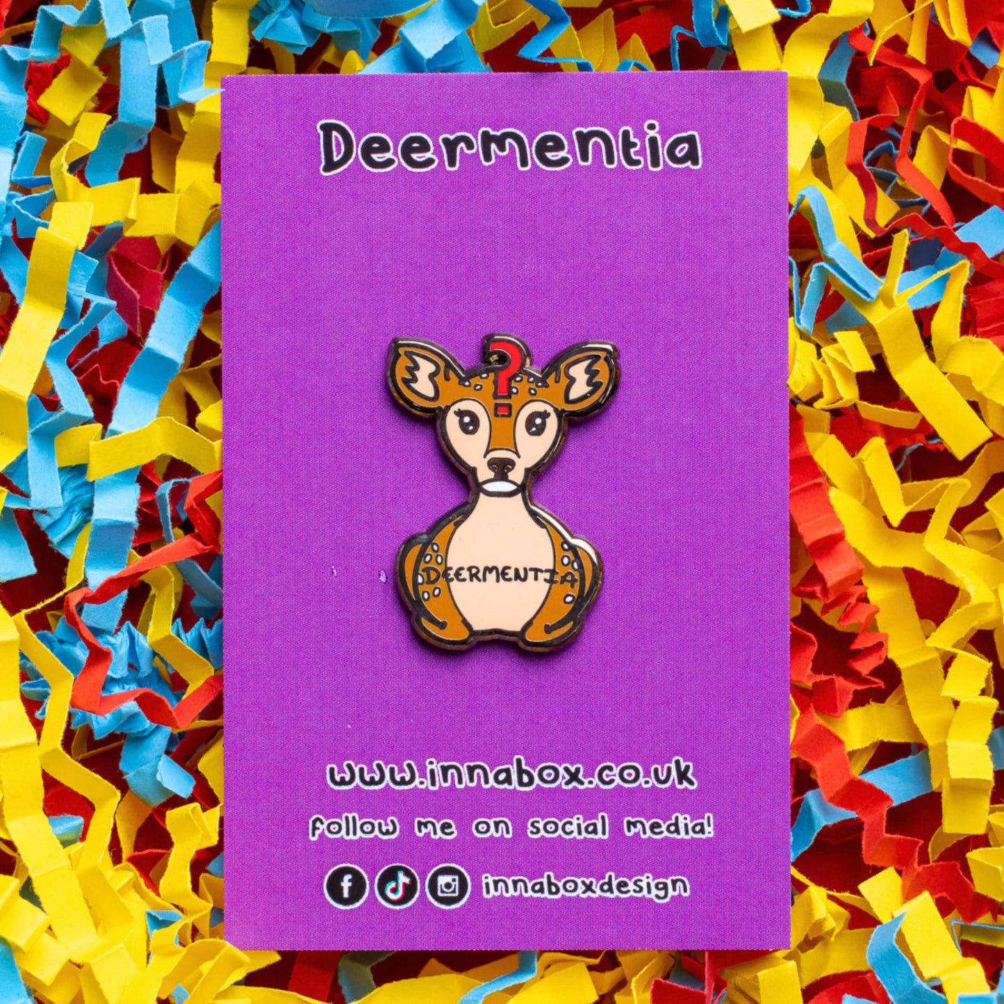 The Deermentia Deer Enamel Pin - Dementia on purple backing card laid on a red, blue and yellow card confetti background. A brown female deer laying down with a vacant expression and red question mark above its head with 'deermentia' written across its middle. The hand drawn design is raising awareness for dementia.