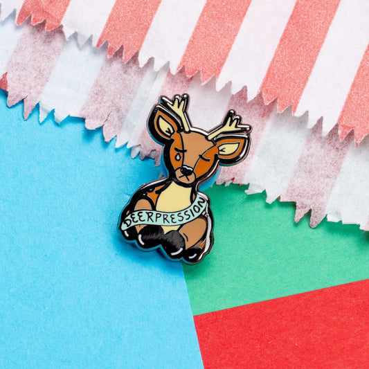 The Deerpression Enamel Pin - Depression on a red, blue and green card background. The deer shape pin its eyes closed with a blue tear falling from one eye, across its front is a blue banner with black text reading 'deerpression'. The design was created to raise awareness for depression.