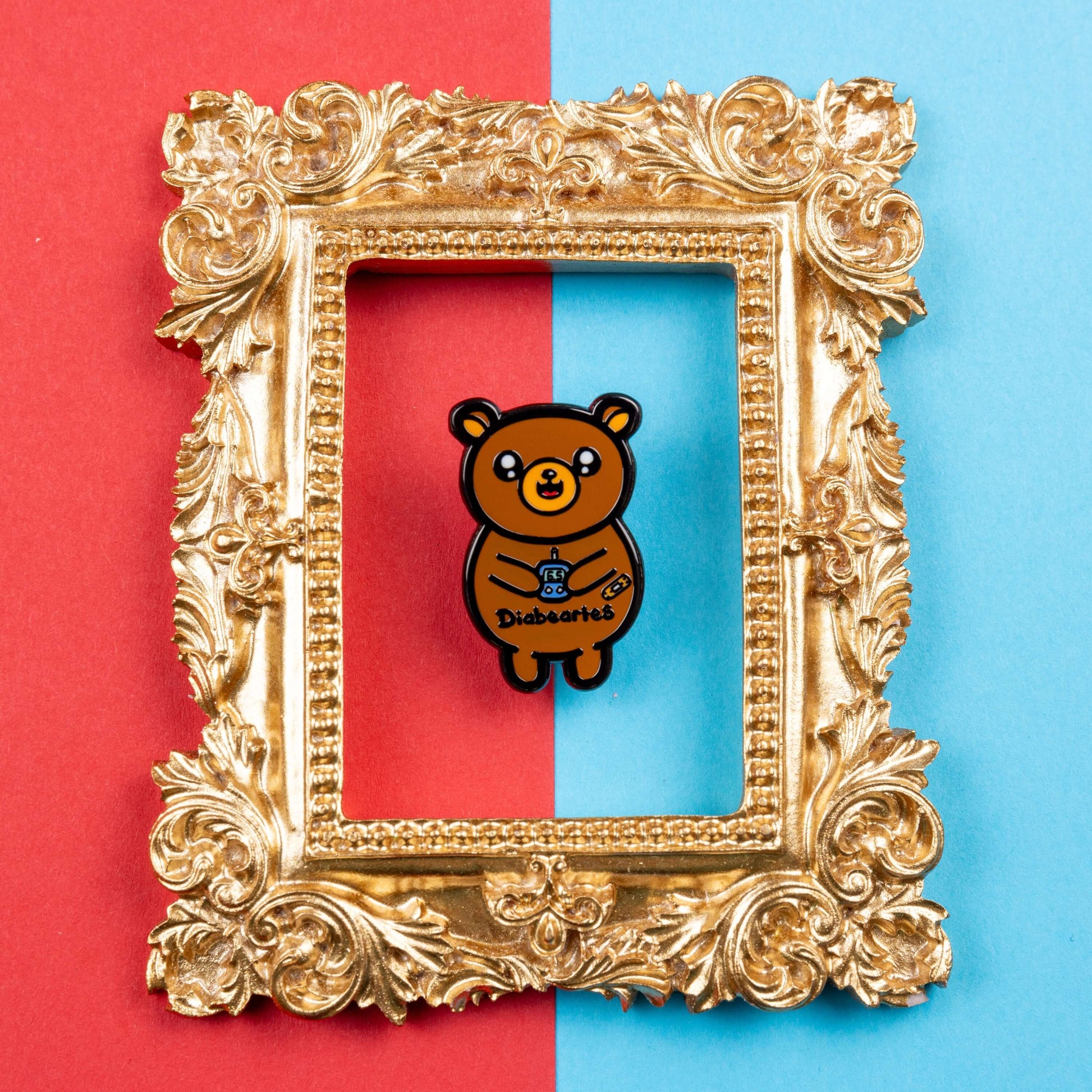 The Diabeartes Enamel Pin - Diabetes on red and blue card inside a gold ornate frame. The brown bear shaped pin is smiling holding a blood glucose reader with a plaster on its arm, across its tummy reads 'diabeartes' in black. The design is raising awareness for those with type 1 diabetes and type 2 diabetes.