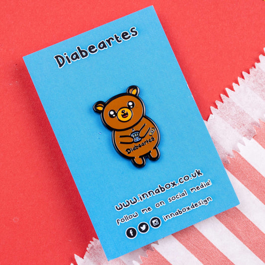 The Diabeartes Enamel Pin - Diabetes on blue backing card laid on red card. The brown bear shaped pin is smiling holding a blood glucose reader with a plaster on its arm, across its tummy reads 'diabeartes' in black. The design is raising awareness for those with type 1 diabetes and type 2 diabetes.