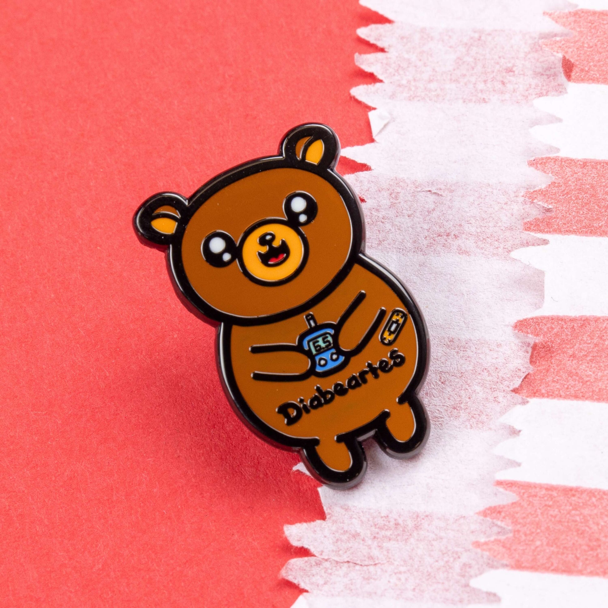 The Diabeartes Enamel Pin - Diabetes on red card. The brown bear shaped pin is smiling holding a blood glucose reader with a plaster on its arm, across its tummy reads 'diabeartes' in black. The design is raising awareness for those with type 1 diabetes and type 2 diabetes.