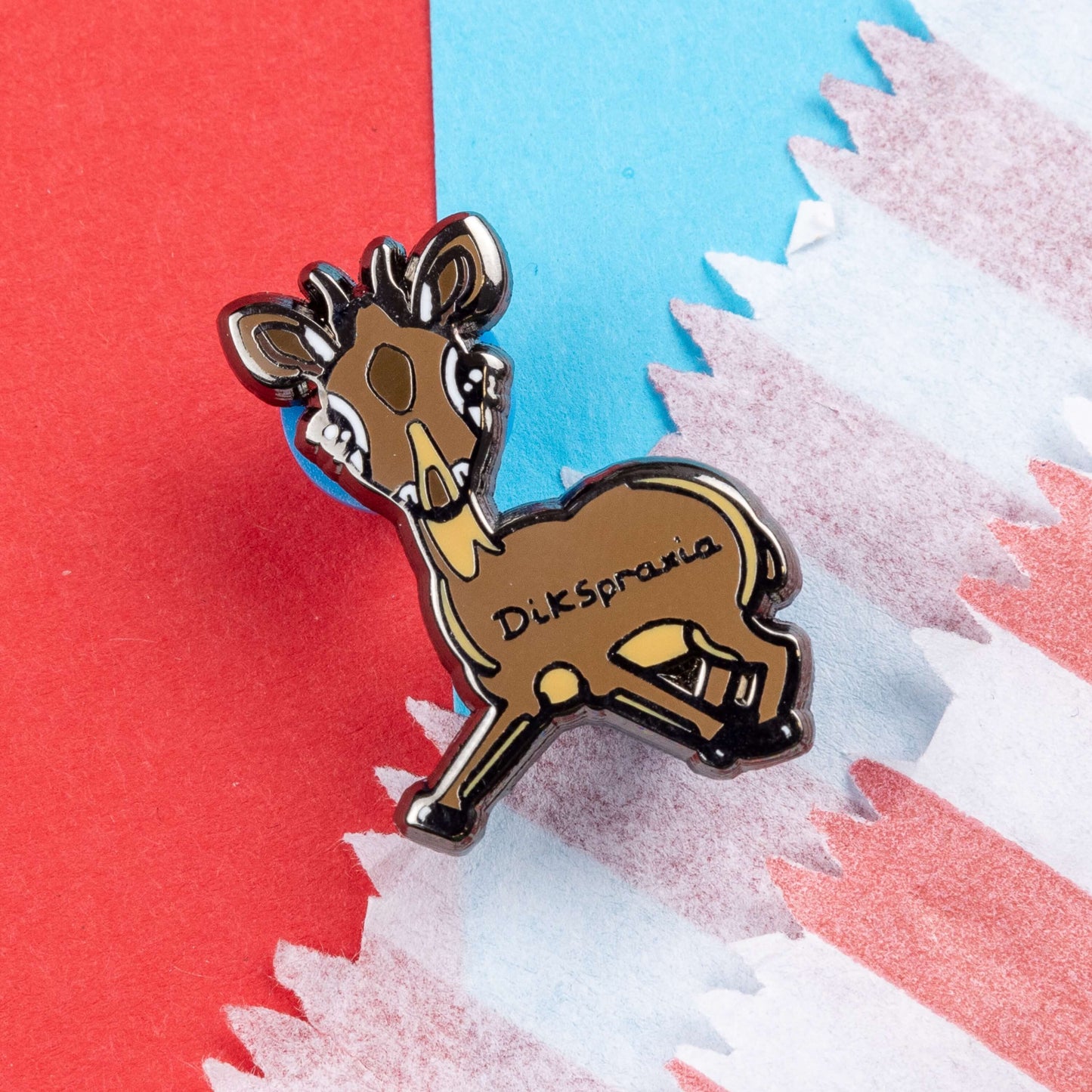 The Dikspraxia Enamel Pin - Dyspraxia on red and blue card. The brown dik dik antelope shaped enamel pin has its two front legs splayed chaotically with black text reading 'dikspraxia' across its middle. The design is raising awareness for dyspraxia and neurodivergence.
