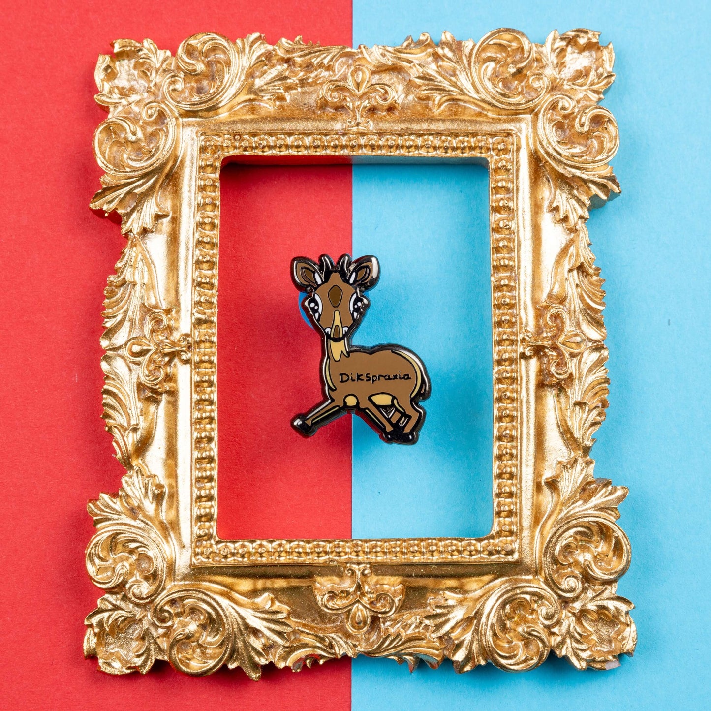 The Dikspraxia Enamel Pin - Dyspraxia on red and blue card in a gold ornate frame. The brown dik dik antelope shaped enamel pin has its two front legs splayed chaotically with black text reading 'dikspraxia' across its middle. The design is raising awareness for dyspraxia and neurodivergence.