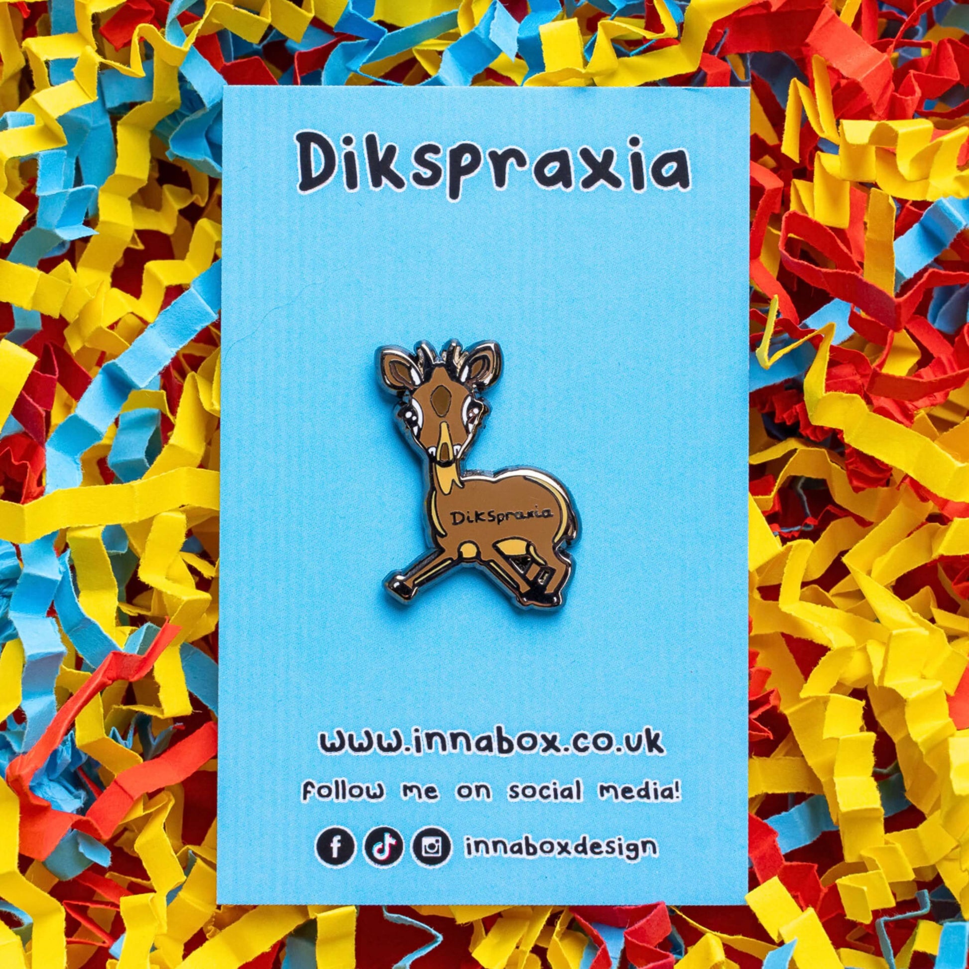The Dikspraxia Enamel Pin - Dyspraxia on blue backing card laid on red, yellow and blue card confetti background. The brown dik dik antelope shaped enamel pin has its two front legs splayed chaotically with black text reading 'dikspraxia' across its middle. The design is raising awareness for dyspraxia and neurodivergence.