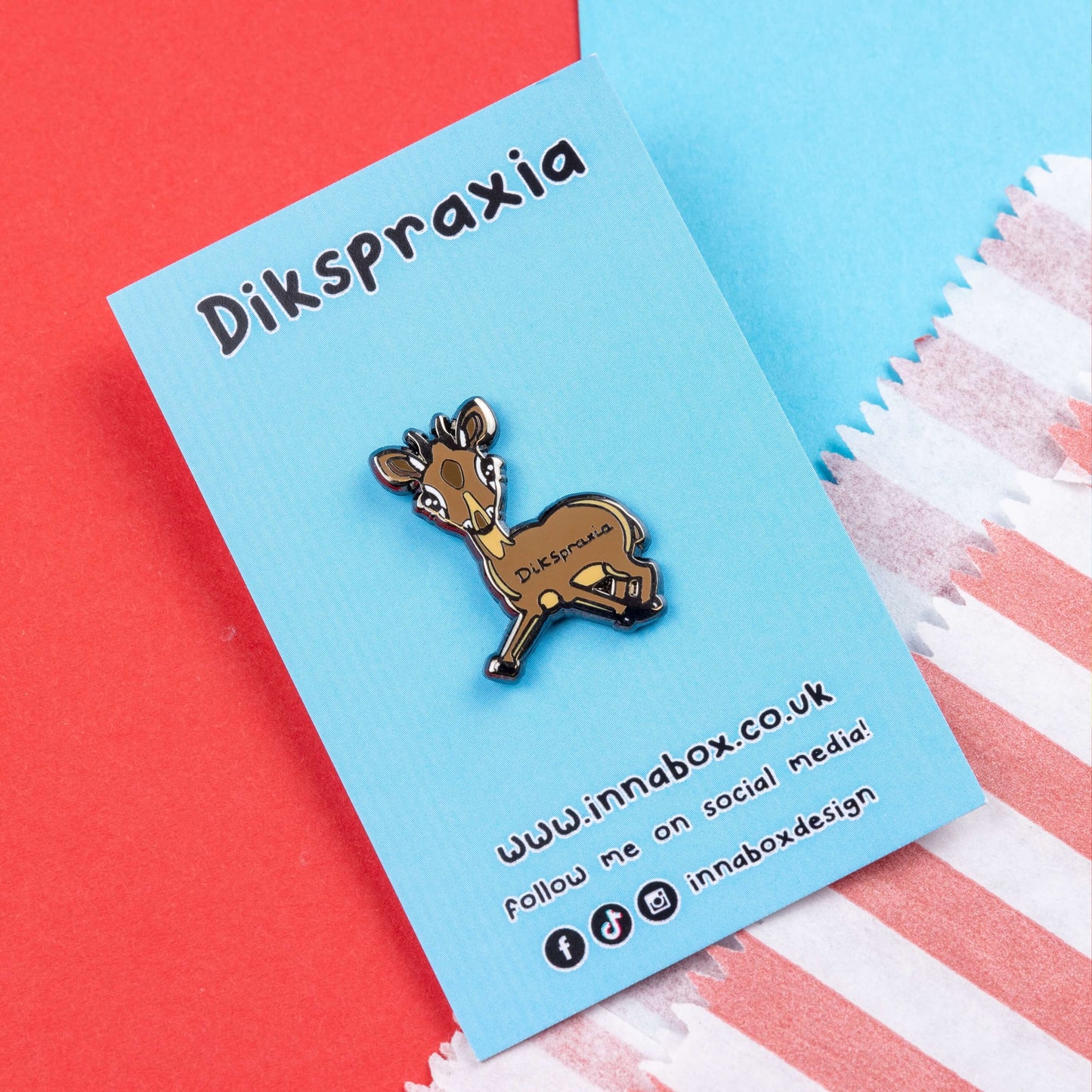 The Dikspraxia Enamel Pin - Dyspraxia on blue backing card laid on red and blue card. The brown dik dik antelope shaped enamel pin has its two front legs splayed chaotically with black text reading 'dikspraxia' across its middle. The design is raising awareness for dyspraxia and neurodivergence.