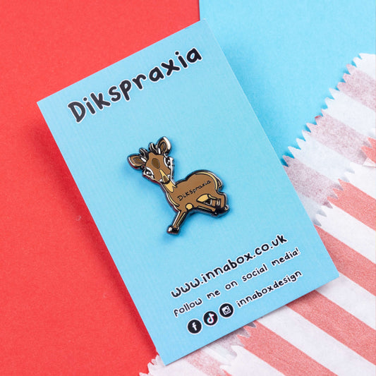 The Dikspraxia Enamel Pin - Dyspraxia on blue backing card laid on red and blue card. The brown dik dik antelope shaped enamel pin has its two front legs splayed chaotically with black text reading 'dikspraxia' across its middle. The design is raising awareness for dyspraxia and neurodivergence.