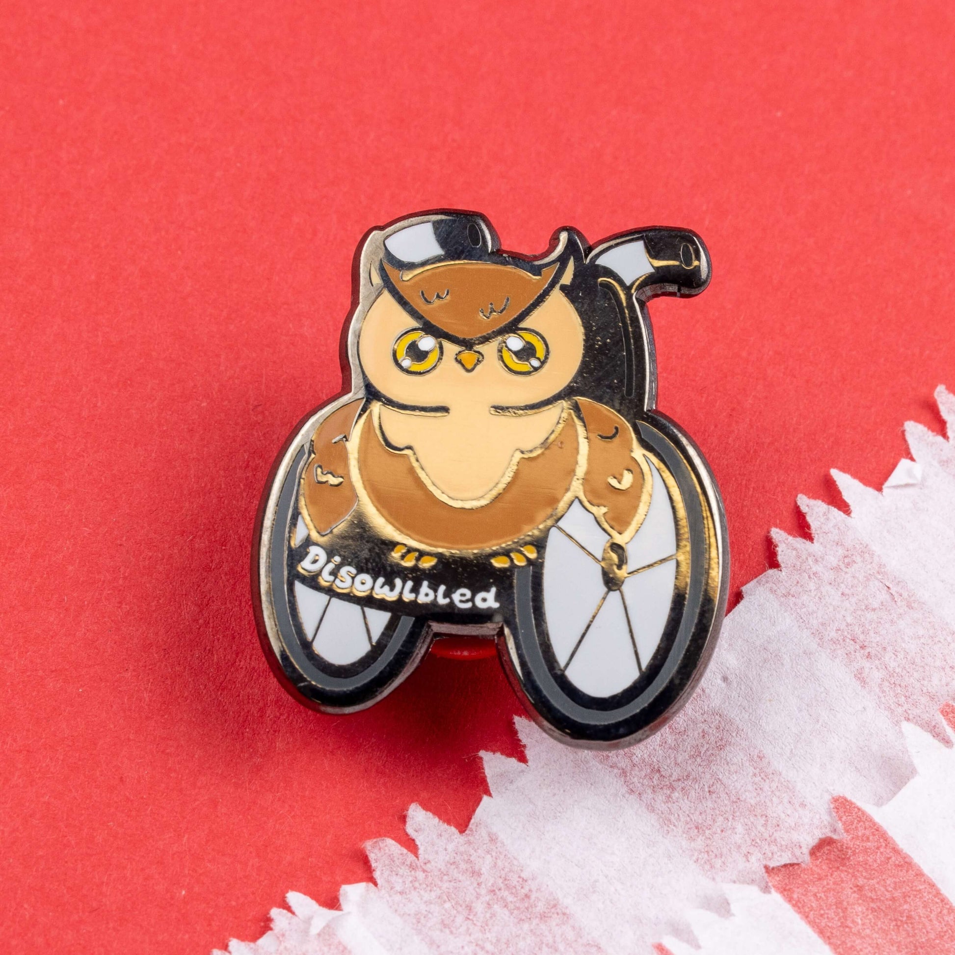 The Disowlbled Enamel Pin - Disabled on red card. A brown tawny owl with yellow eyes sat on a silver and black wheelchair that has white text reading 'disowlbled'. The design is raising awareness for disabilities and invisible illnesses.