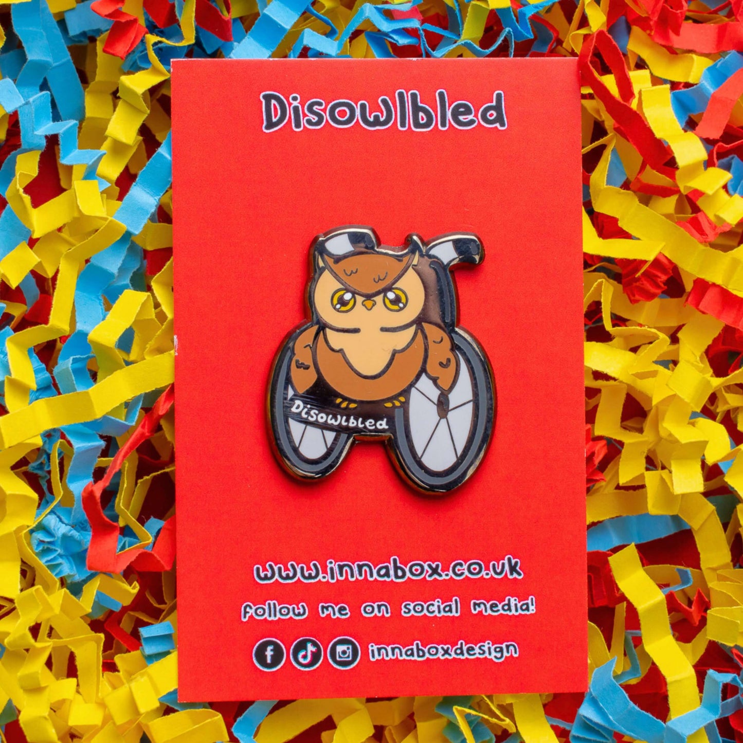 The Disowlbled Enamel Pin - Disabled on red backing card on red card laid on a red, yellow & blue card confetti background. A brown tawny owl with yellow eyes sat on a silver and black wheelchair that has white text reading 'disowlbled'. The design is raising awareness for disabilities and invisible illnesses.