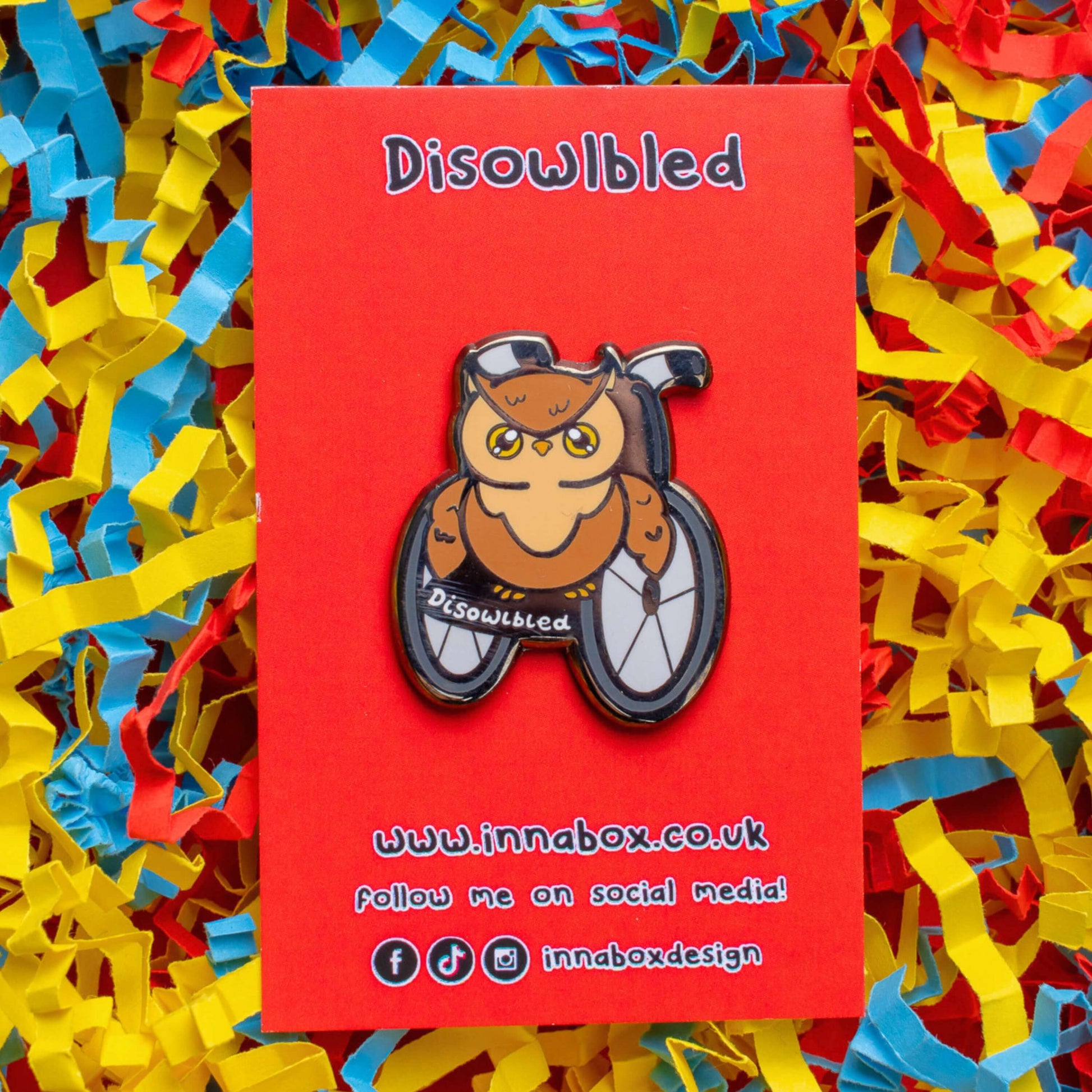 The Disowlbled Enamel Pin - Disabled on red backing card on red card laid on a red, yellow & blue card confetti background. A brown tawny owl with yellow eyes sat on a silver and black wheelchair that has white text reading 'disowlbled'. The design is raising awareness for disabilities and invisible illnesses.