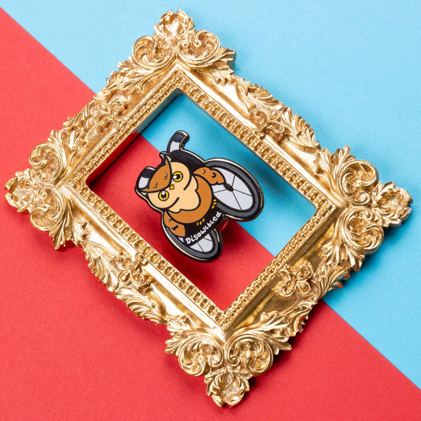 The Disowlbled Enamel Pin - Disabled on red and blue card inside a gold ornate frame. A brown tawny owl with yellow eyes sat on a silver and black wheelchair that has white text reading 'disowlbled'. The design is raising awareness for disabilities and invisible illnesses.