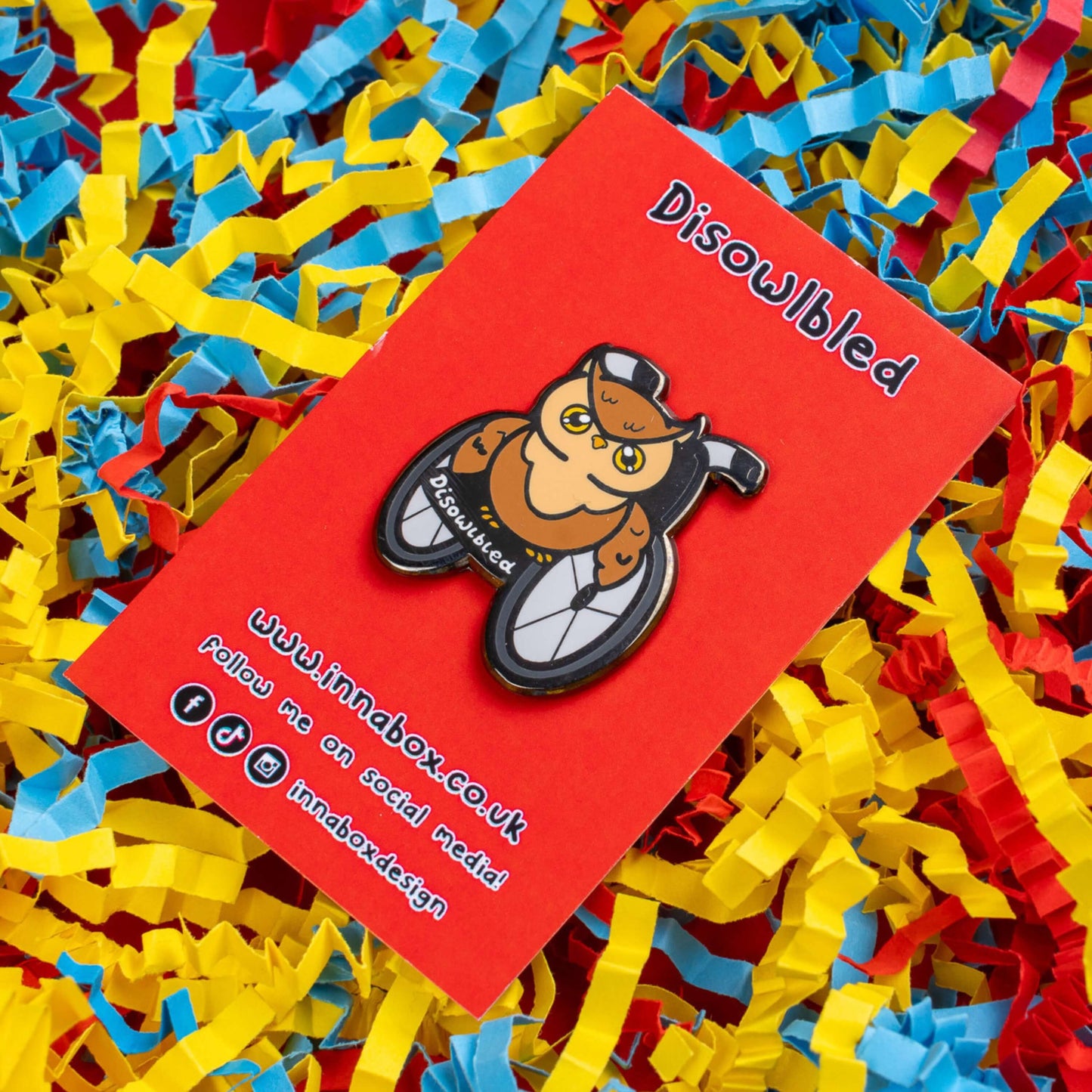 The Disowlbled Enamel Pin - Disabled on red backing card on red card laid on a red, yellow & blue card confetti background. A brown tawny owl with yellow eyes sat on a silver and black wheelchair that has white text reading 'disowlbled'. The design is raising awareness for disabilities and invisible illnesses.