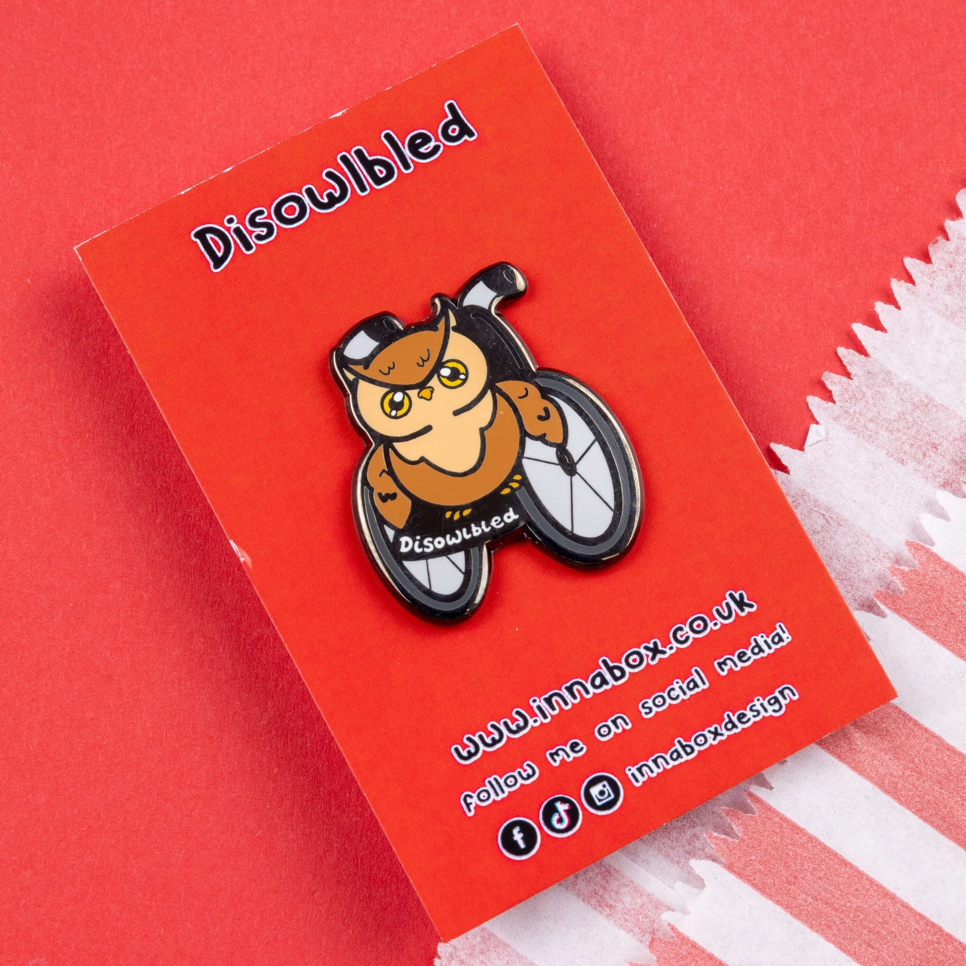 The Disowlbled Enamel Pin - Disabled on red backing card on red card. A brown tawny owl with yellow eyes sat on a silver and black wheelchair that has white text reading 'disowlbled'. The design is raising awareness for disabilities and invisible illnesses.