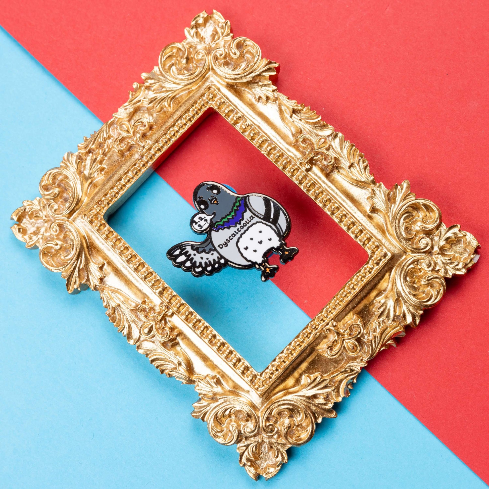 The Dyscalcoolia Pigeon Enamel Pin - Dyscalculia on a red and blue background in a gold ornate frame. A Pigeon looking confused holding up a wing with a speech bubble full of numbers and a question mark across its chest reads 'dyscalcoolia'. The enamel pin is raising awareness for dyscalculia.