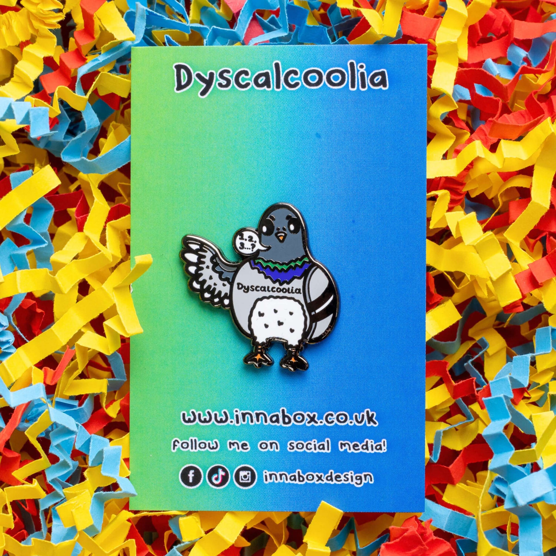 The Dyscalcoolia Pigeon Enamel Pin - Dyscalculia on a blue to green gradient backing card with black text laid on a red, yellow and blue confetti card background. A Pigeon looking confused holding up a wing with a speech bubble full of numbers and a question mark across its chest reads 'dyscalcoolia'. The enamel pin is raising awareness for dyscalculia.