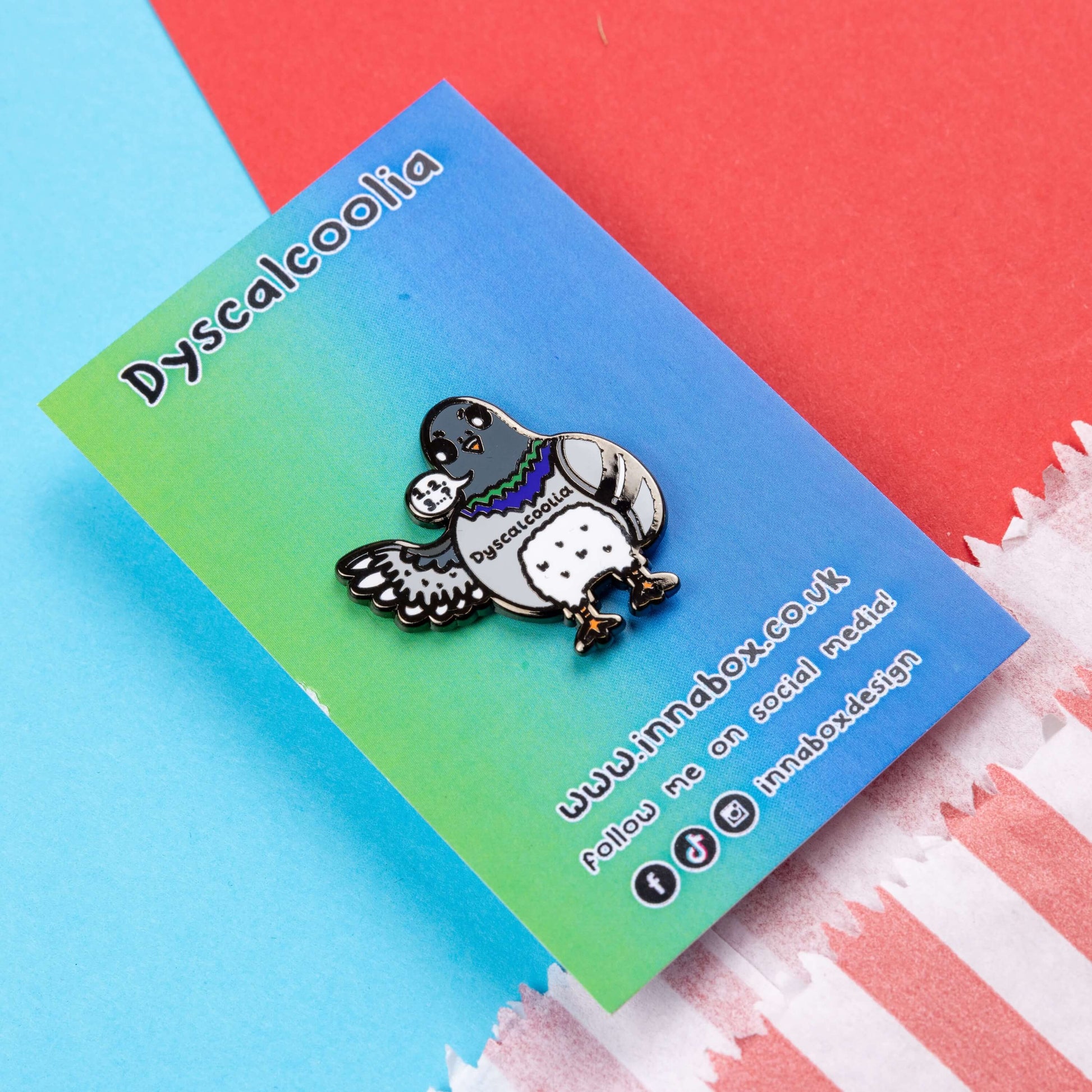 The Dyscalcoolia Pigeon Enamel Pin - Dyscalculia on a blue to green gradient backing card with black text. A Pigeon looking confused holding up a wing with a speech bubble full of numbers and a question mark across its chest reads 'dyscalcoolia'. The enamel pin is raising awareness for dyscalculia.