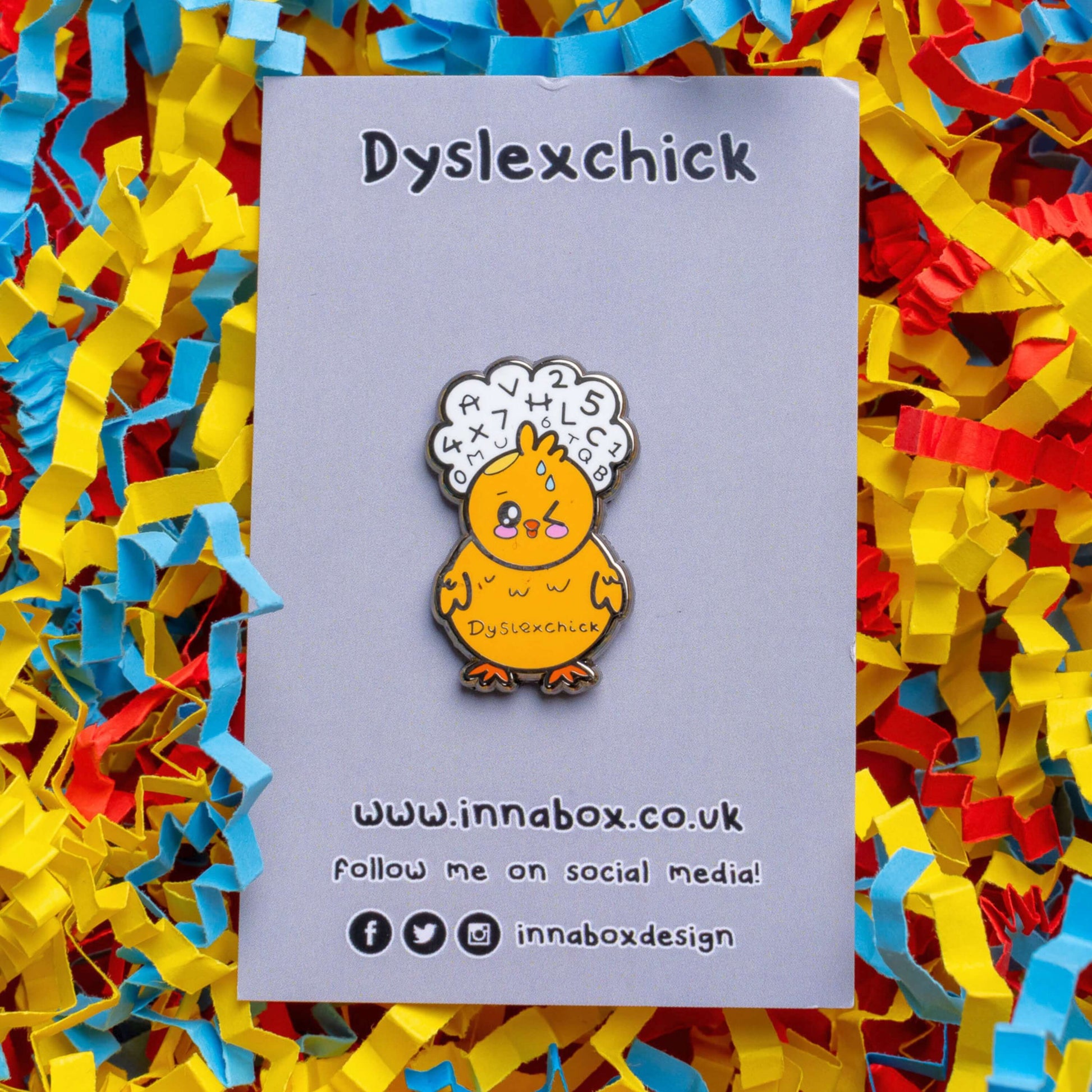 The Dyslexchick Enamel Pin - Dyslexia on grey backing card laid on a red, yellow and blue card confetti background. A yellow confused chick shaped pin badge with a thought bubble above its head full of letters and numbers with 'dyslexchick' written across its middle. The hand drawn design is raising awareness for dyslexia.