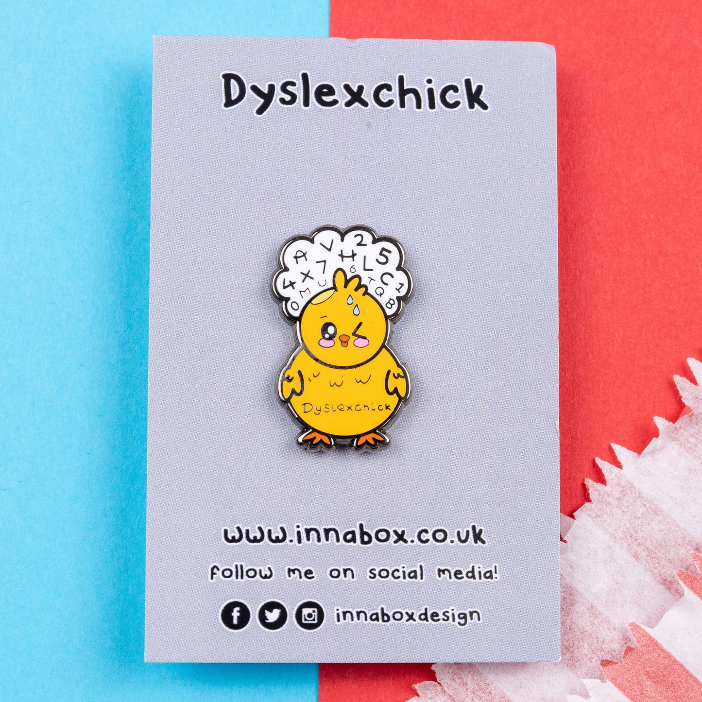 The Dyslexchick Enamel Pin - Dyslexia on grey backing card laid on a red and blue background. A yellow confused chick shaped pin badge with a thought bubble above its head full of letters and numbers with 'dyslexchick' written across its middle. The hand drawn design is raising awareness for dyslexia.