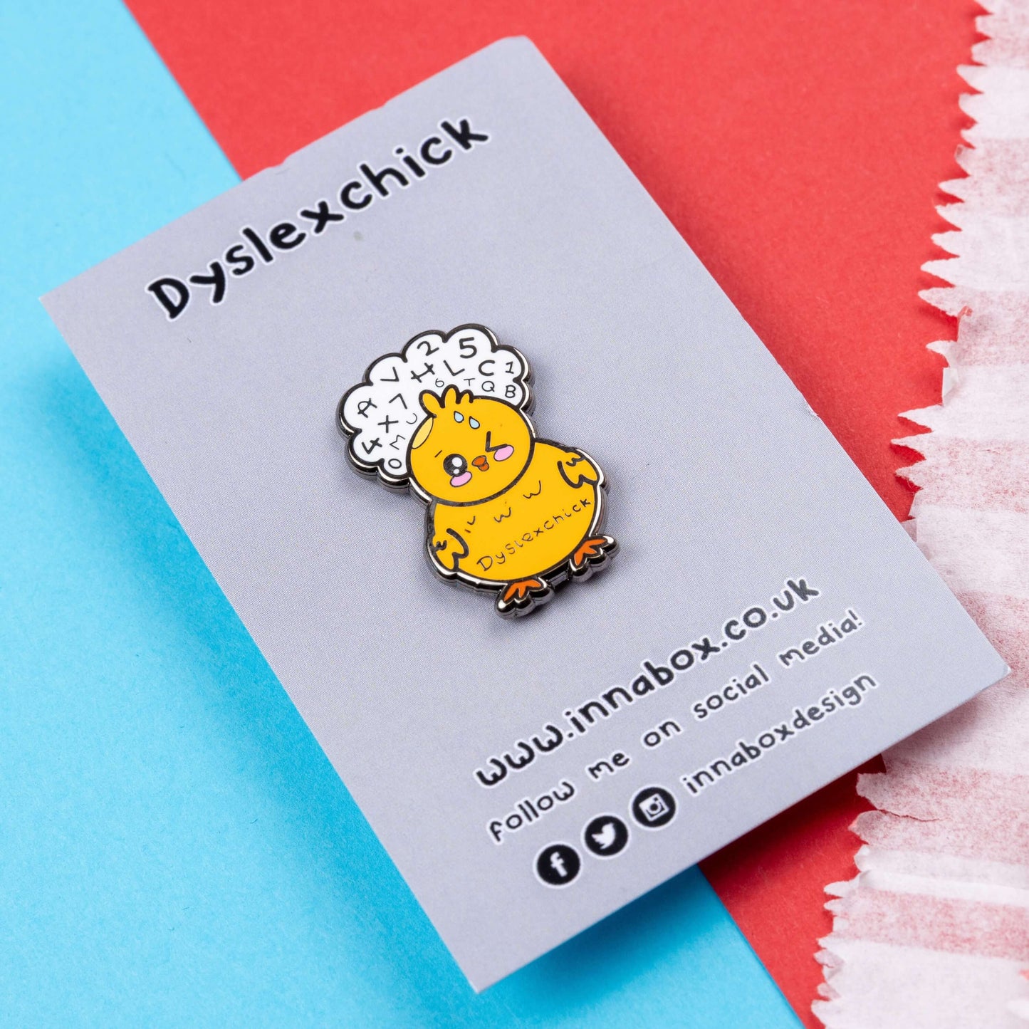 The Dyslexchick Enamel Pin - Dyslexia on grey backing card laid on a red and blue background. A yellow confused chick shaped pin badge with a thought bubble above its head full of letters and numbers with 'dyslexchick' written across its middle. The hand drawn design is raising awareness for dyslexia.
