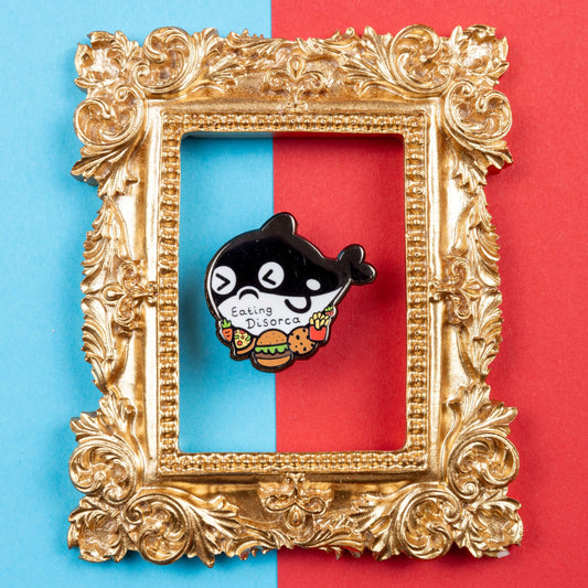 The Eating Disorca Orca Whale Enamel Pin - Eating Disorder on a blue and red background in a gold ornate frame. The black orca whale shaped enamel pin has a stressed expression whilst surrounded by burgers, pizzas, cookies, fries and fruit with 'eating discorca' across its middle. The design is raising awareness for eating disorders.