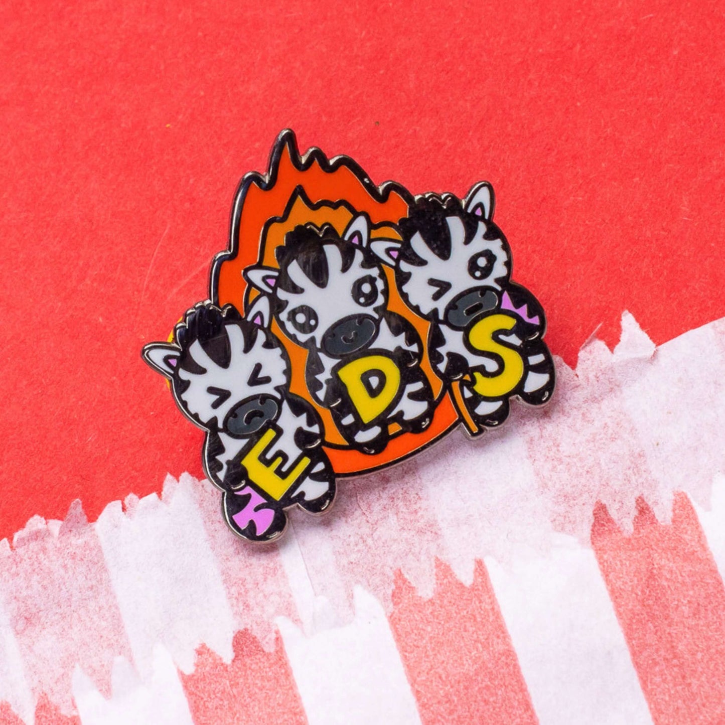 The Ehlers Dazzle Syndrome EDS Enamel Pin 2.0 - Ehlers-Danlos Syndrome on a red card background. A pin of three zebras in front of an orange flame holding up yellow letters spelling out EDS, the first zebra has a swollen pink leg, the next is smiling and the third has a swollen pink arm and a walking stick. Raising awareness for Ehlers-Danlos Syndrome.