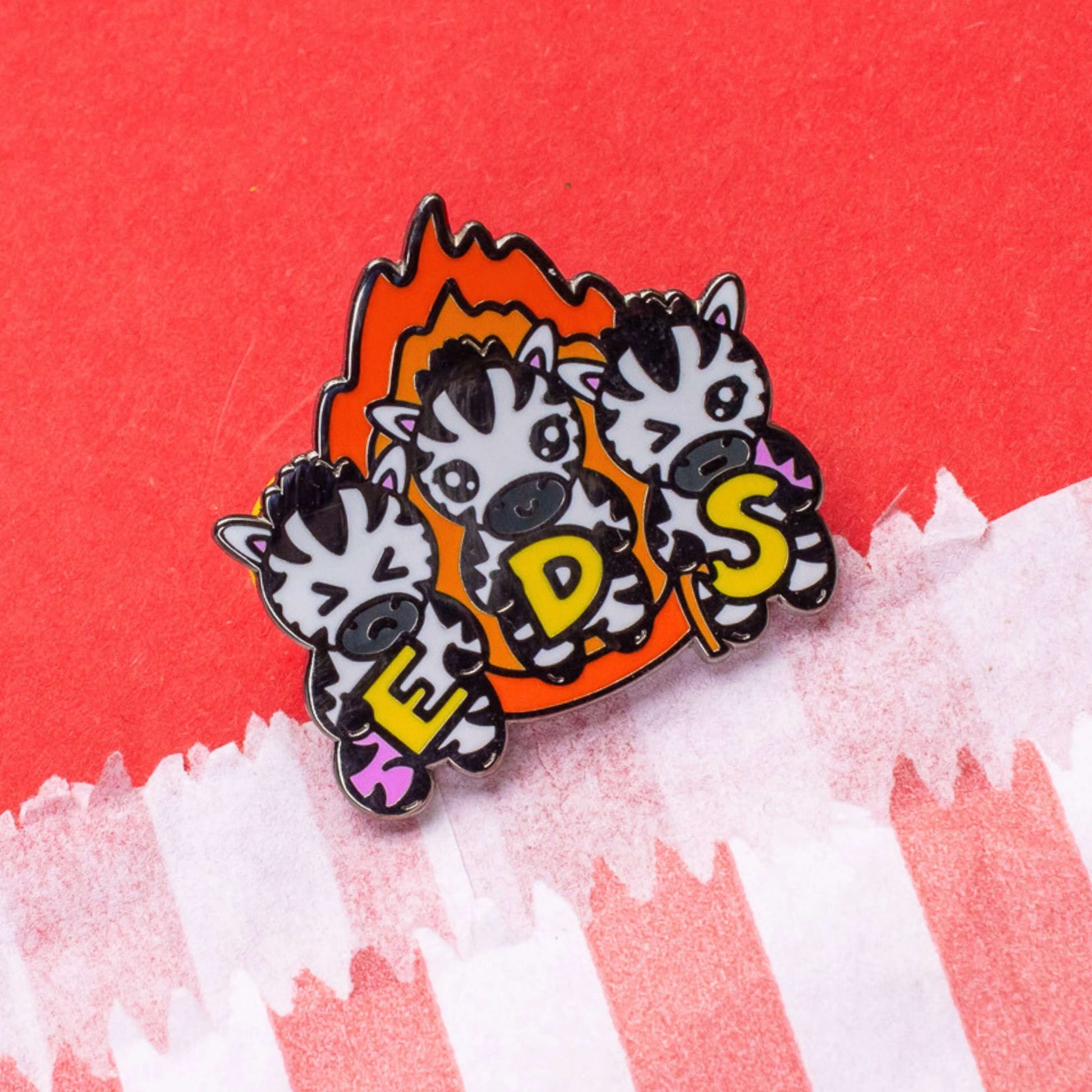 The Ehlers Dazzle Syndrome EDS Enamel Pin 2.0 - Ehlers-Danlos Syndrome on a red card background. A pin of three zebras in front of an orange flame holding up yellow letters spelling out EDS, the first zebra has a swollen pink leg, the next is smiling and the third has a swollen pink arm and a walking stick. Raising awareness for Ehlers-Danlos Syndrome.