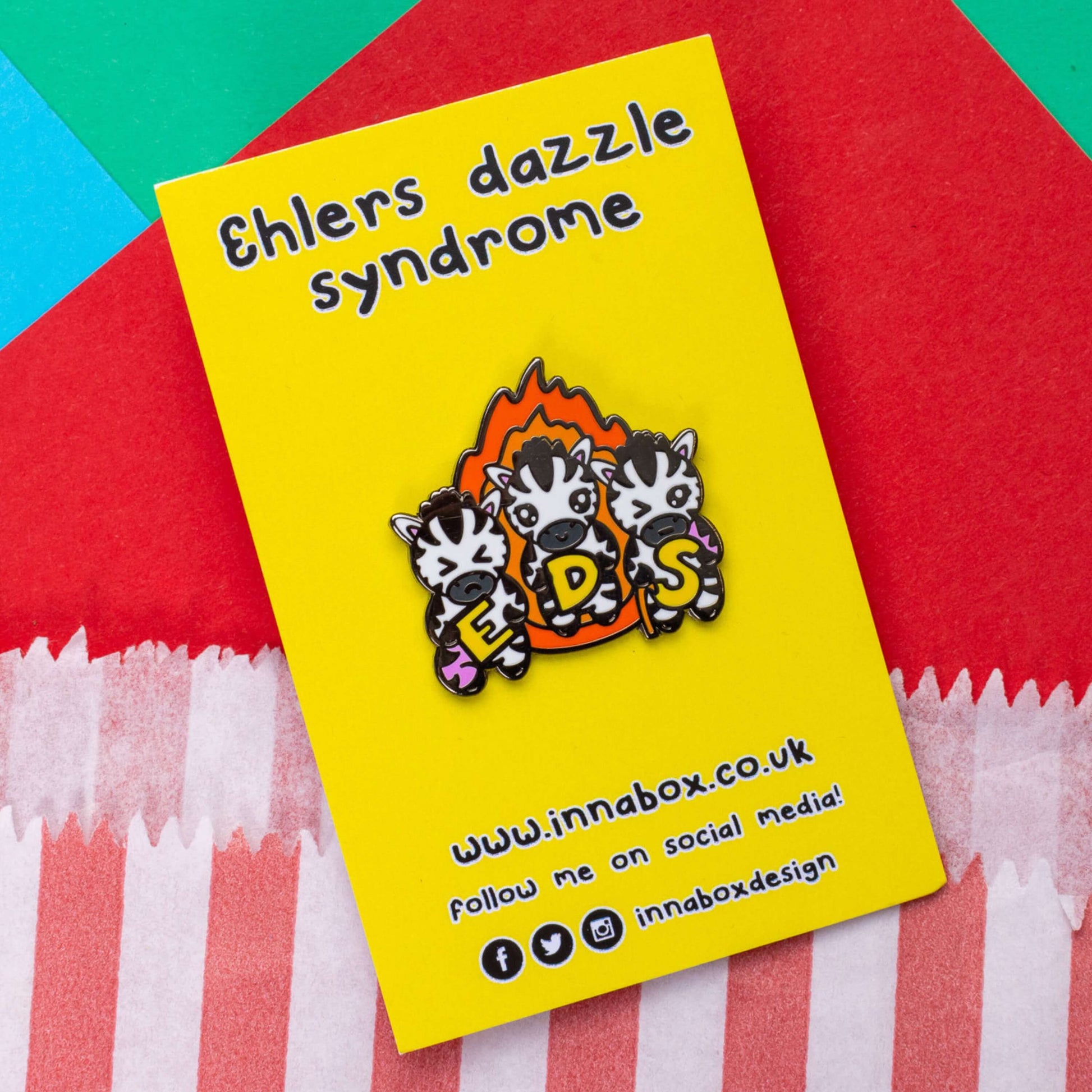 The Ehlers Dazzle Syndrome EDS Enamel Pin 2.0 - Ehlers-Danlos Syndrome on yellow backing card with black text above reading 'ehlers dazzle syndrome' and below reading the innabox website and social media handles on red, green and blue card. A pin of three zebras in front of an orange flame holding up yellow letters spelling out EDS, the first zebra has a swollen pink leg, the next is smiling and the third has a swollen pink arm and a walking stick. Raising awareness for Ehlers-Danlos Syndrome.