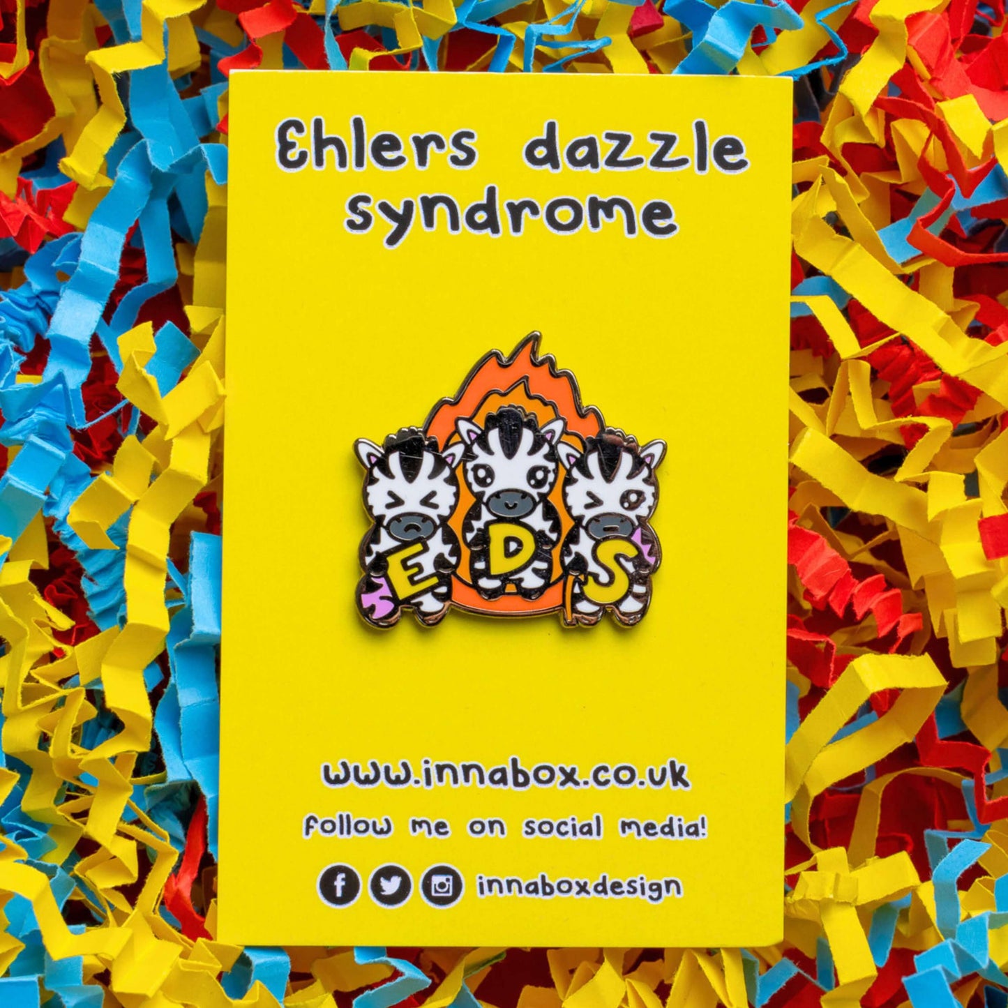 The Ehlers Dazzle Syndrome EDS Enamel Pin 2.0 - Ehlers-Danlos Syndrome on yellow backing card with black text above reading 'ehlers dazzle syndrome' and below reading the innabox website and social media handles on red, yellow and blue card confetti. A pin of three zebras in front of an orange flame holding up yellow letters spelling out EDS, the first zebra has a swollen pink leg, the next is smiling and the third has a swollen pink arm and a walking stick. Raising awareness for Ehlers-Danlos Syndrome.