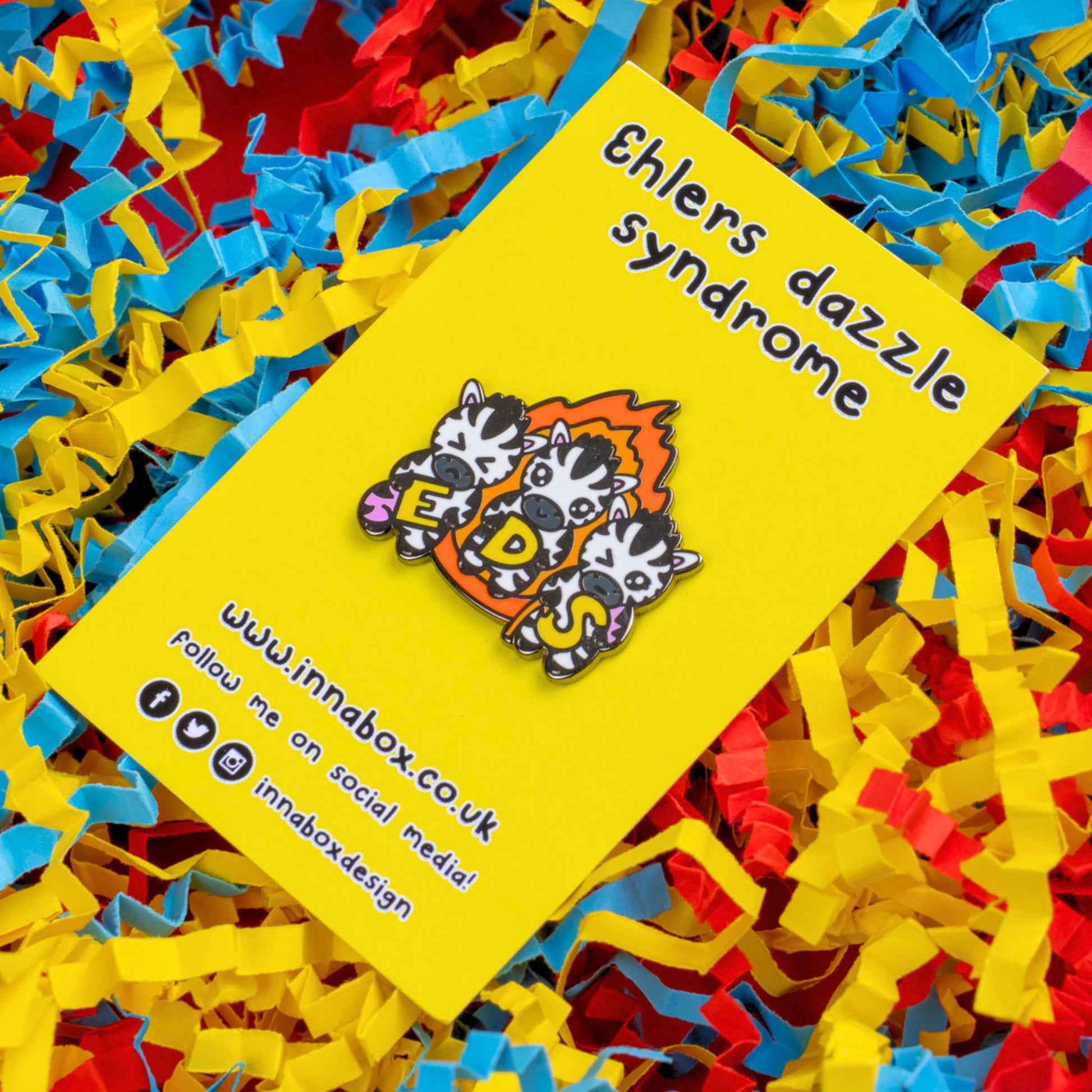 The Ehlers Dazzle Syndrome EDS Enamel Pin 2.0 - Ehlers-Danlos Syndrome on yellow backing card with black text above reading 'ehlers dazzle syndrome' and below reading the innabox website and social media handles on red, yellow and blue card confetti. A pin of three zebras in front of an orange flame holding up yellow letters spelling out EDS, the first zebra has a swollen pink leg, the next is smiling and the third has a swollen pink arm and a walking stick. Raising awareness for Ehlers-Danlos Syndrome.