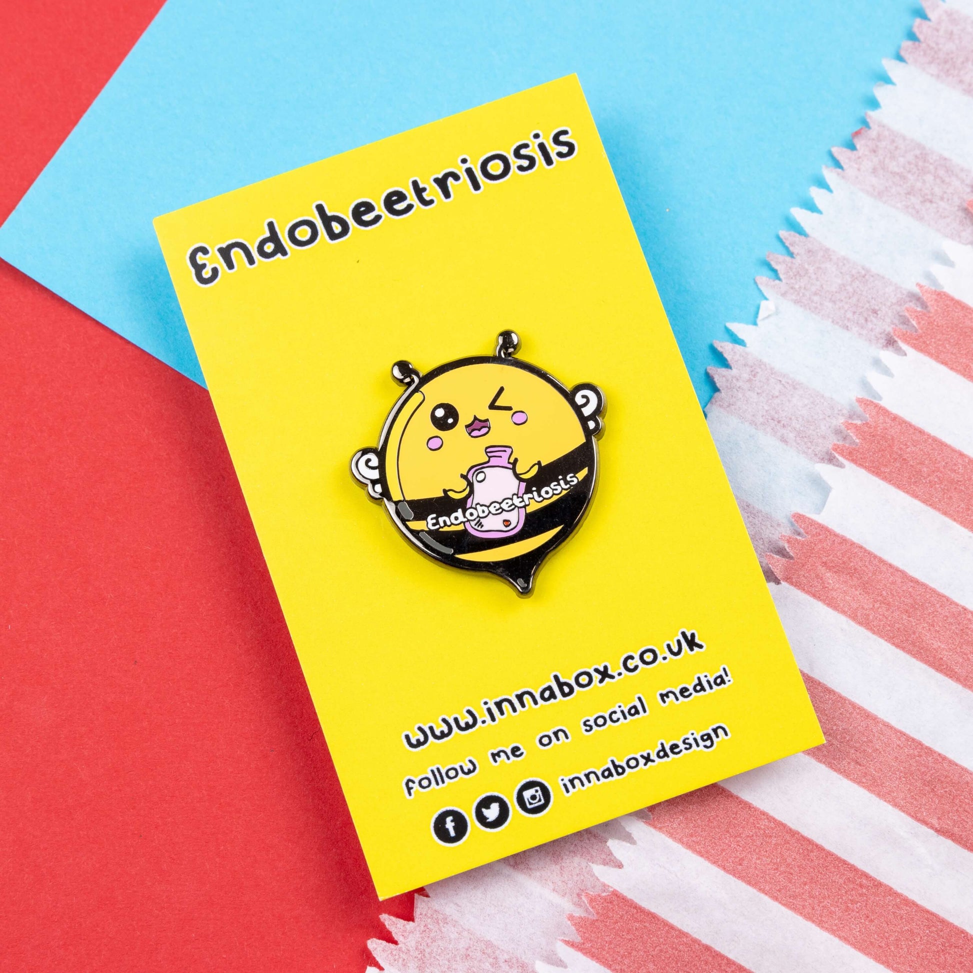 Endobeetriosis 2.0 Bee Enamel Pin - Endometriosis on yellow backing card laid on a red and blue card background. The enamel pin is a bumble bee with a smiley face and holding a pink hot waterbottle in its hands, across its middle is white text that reads endobeetriosis. The enamel pin is designed to raise awareness for endometriosis.