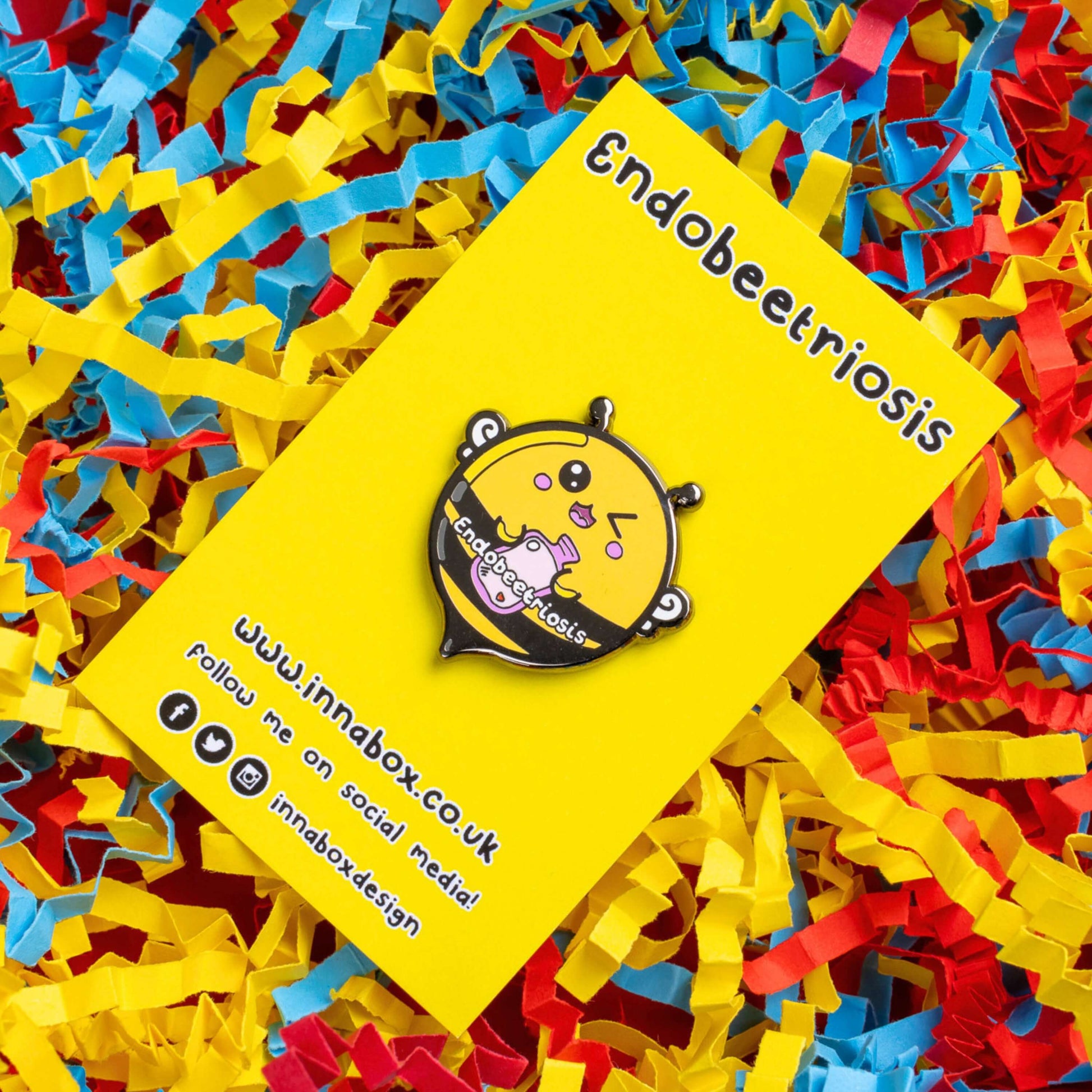 Endobeetriosis 2.0 Bee Enamel Pin - Endometriosis on yellow backing card laid on a yellow, red and blue card confetti background. The enamel pin is a bumble bee with a smiley face and holding a pink hot waterbottle in its hands, across its middle is white text that reads endobeetriosis. The enamel pin is designed to raise awareness for endometriosis.