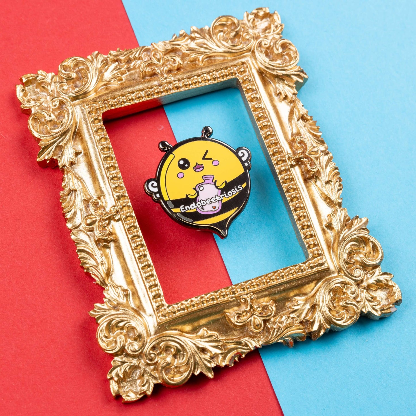 Endobeetriosis 2.0 Bee Enamel Pin - Endometriosis on a red and blue card background in a gold ornate frame. The enamel pin is a bumble bee with a smiley face and holding a pink hot waterbottle in its hands, across its middle is white text that reads endobeetriosis. The enamel pin is designed to raise awareness for endometriosis.