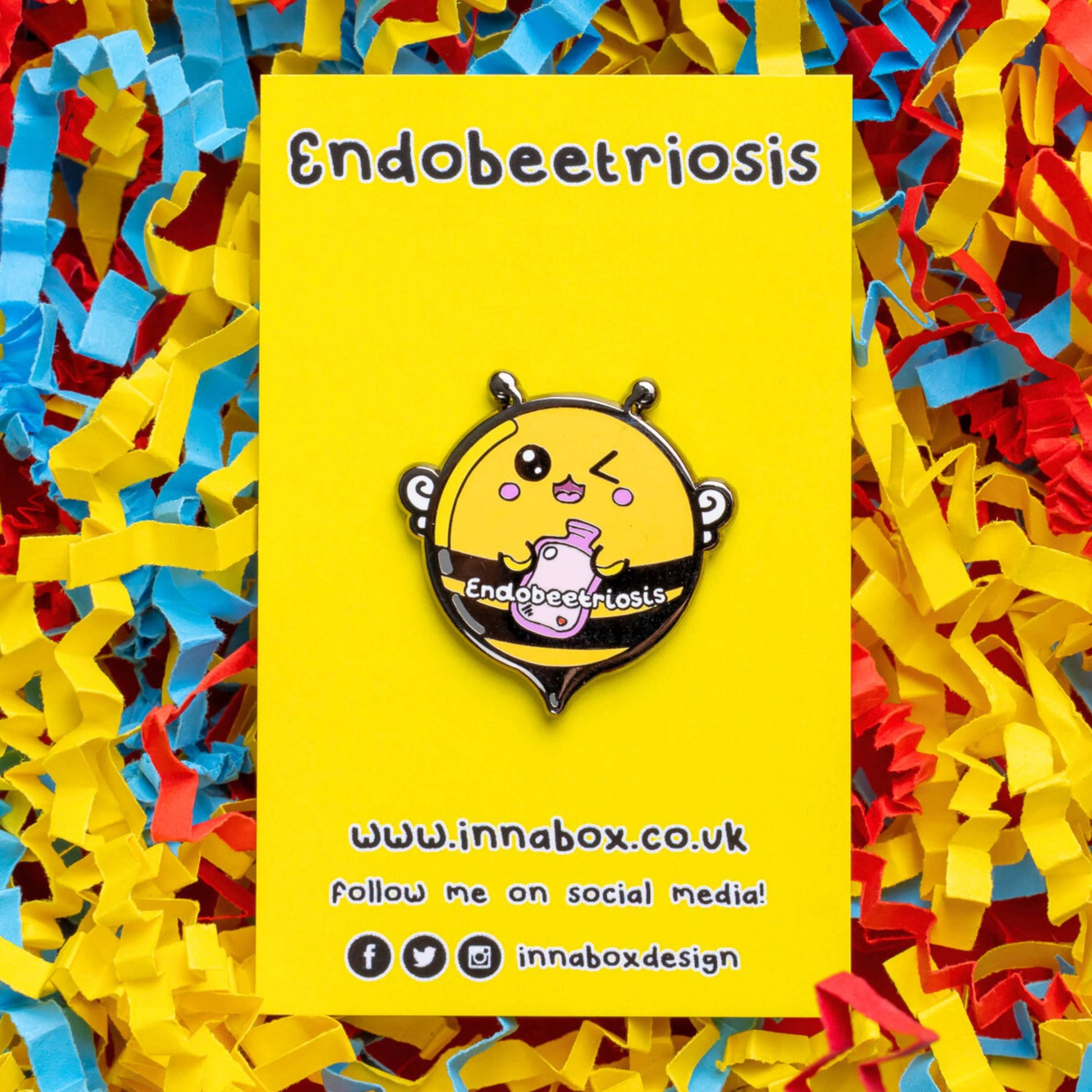 Endobeetriosis 2.0 Bee Enamel Pin - Endometriosis on yellow backing card laid on a yellow, red and blue card confetti background. The enamel pin is a bumble bee with a smiley face and holding a pink hot waterbottle in its hands, across its middle is white text that reads endobeetriosis. The enamel pin is designed to raise awareness for endometriosis.