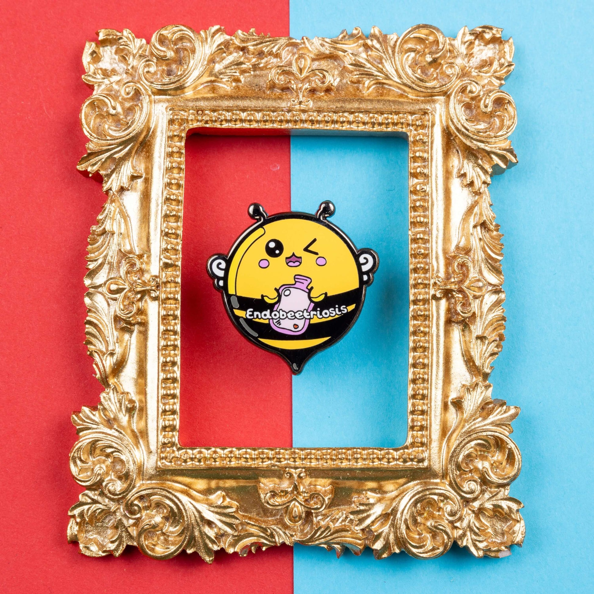 Endobeetriosis 2.0 Bee Enamel Pin - Endometriosis on a red and blue card background in a gold ornate frame. The enamel pin is a bumble bee with a smiley face and holding a pink hot waterbottle in its hands, across its middle is white text that reads endobeetriosis. The enamel pin is designed to raise awareness for endometriosis.