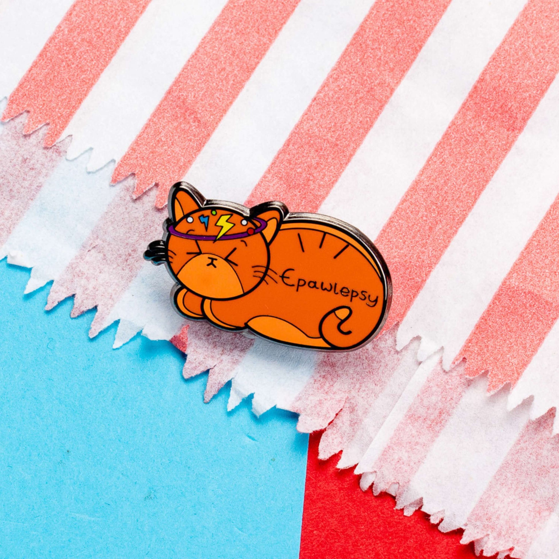 Epawlepsy Enamel Pin - Epilepsy on a red and blue card background. The enamel pin is a ginger cat with eyes scrunched closed and symbols to represent a dizzy spell drawn across his head. Epawlepsy is written on the cats stomach. Enamel pin is designed to raise awareness for epilepsy.