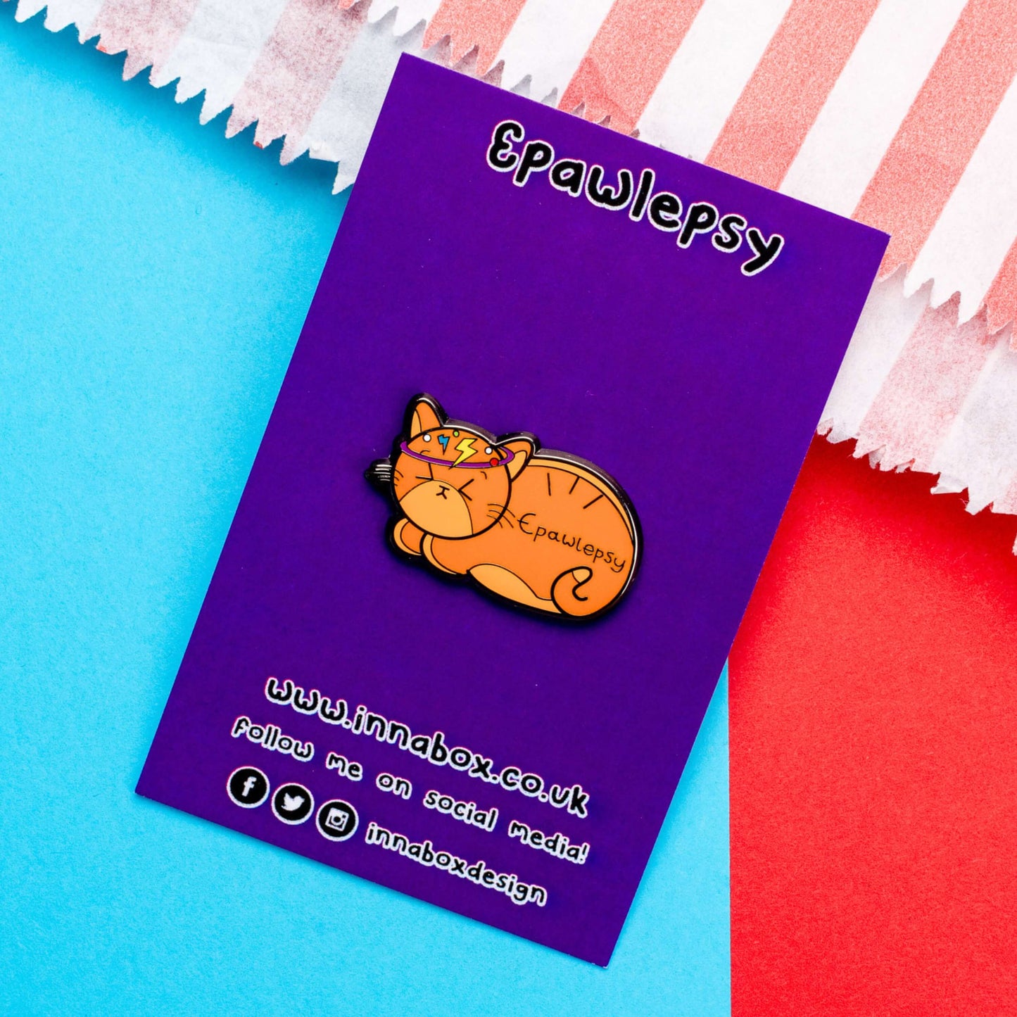 Epawlepsy Enamel Pin - Epilepsy on a purple backing card red background. The enamel pin is a ginger cat with eyes scrunched closed and symbols to represent a dizzy spell drawn across his head. Epawlepsy is written on the cats stomach. Enamel pin is designed to raise awareness for epilepsy.