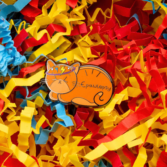 Epawlepsy Enamel Pin - Epilepsy on a red, blue and yellow card confetti background. The enamel pin is a ginger cat with eyes scrunched closed and symbols to represent a dizzy spell drawn across his head. Epawlepsy is written on the cats stomach. Enamel pin is designed to raise awareness for epilepsy.