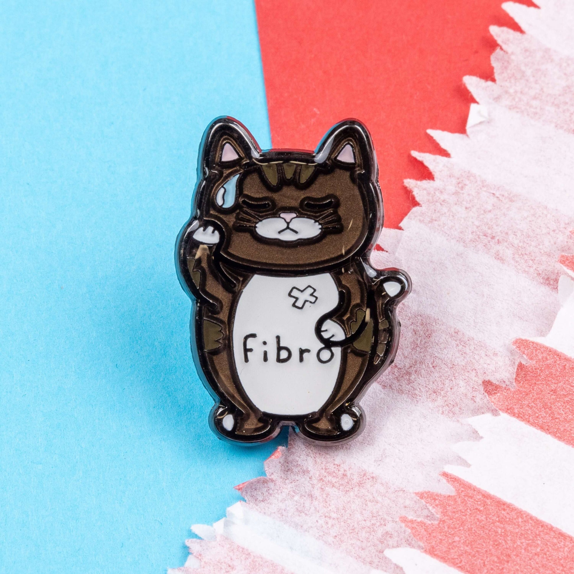 Fibromeowgia Enamel Pin - Fibromyalgia on a red and blue background. The enamel pin is a brown cat with a white stomach with fibro written across it. The cat has its eyes closed, a sweat droplet on its forehead and holding its head in its paw. The enamel pin is designed to raise awareness for fibromyalgia.