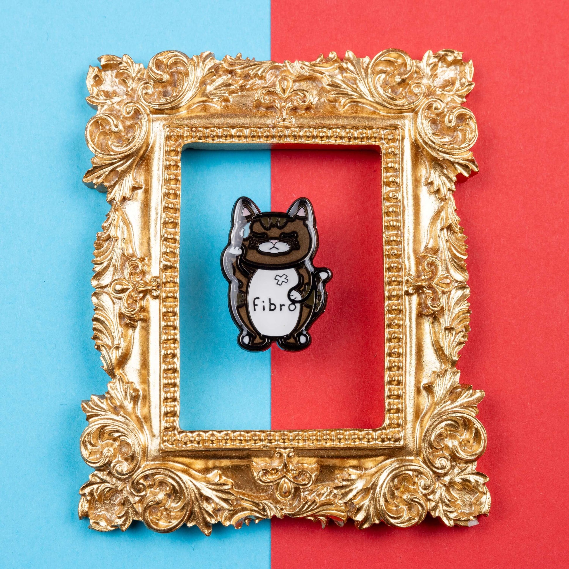 Fibromeowgia Enamel Pin - Fibromyalgia on a red and blue background inside a gold ornate frame. The enamel pin is a brown cat with a white stomach with fibro written across it. The cat has its eyes closed, a sweat droplet on its forehead and holding its head in its paw. The enamel pin is designed to raise awareness for fibromyalgia.