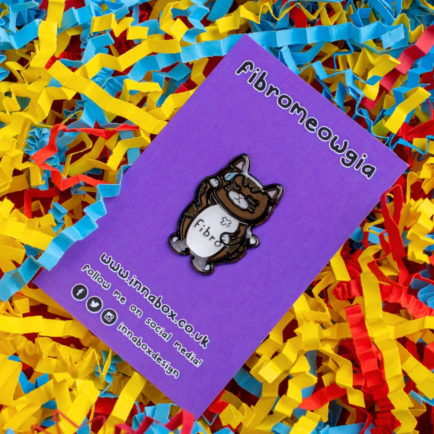 Fibromeowgia Enamel Pin - Fibromyalgia on purple backing card laid on a red, yellow and blue card confetti background. The enamel pin is a brown cat with a white stomach with fibro written across it. The cat has its eyes closed, a sweat droplet on its forehead and holding its head in its paw. The enamel pin is designed to raise awareness for fibromyalgia.