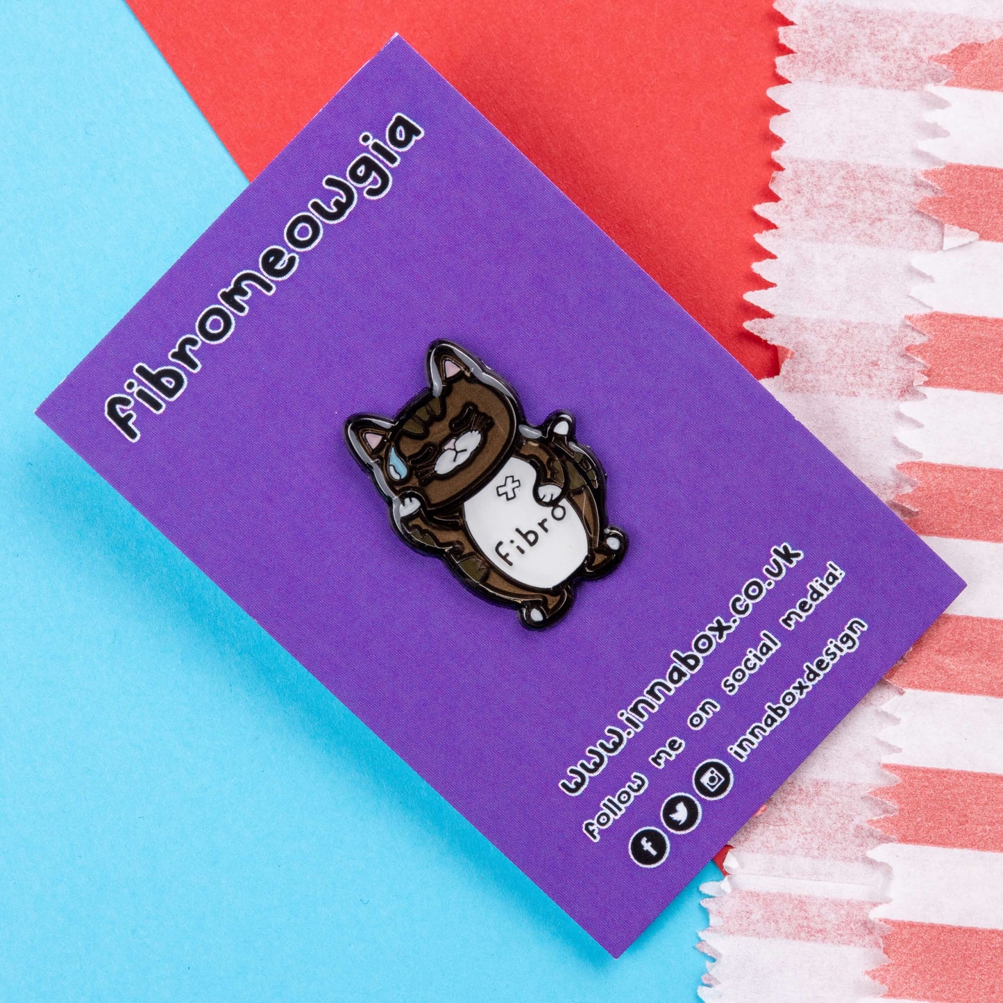 Fibromeowgia Enamel Pin - Fibromyalgia on purple backing card laid on a red and blue background. The enamel pin is a brown cat with a white stomach with fibro written across it. The cat has its eyes closed, a sweat droplet on its forehead and holding its head in its paw. The enamel pin is designed to raise awareness for fibromyalgia.