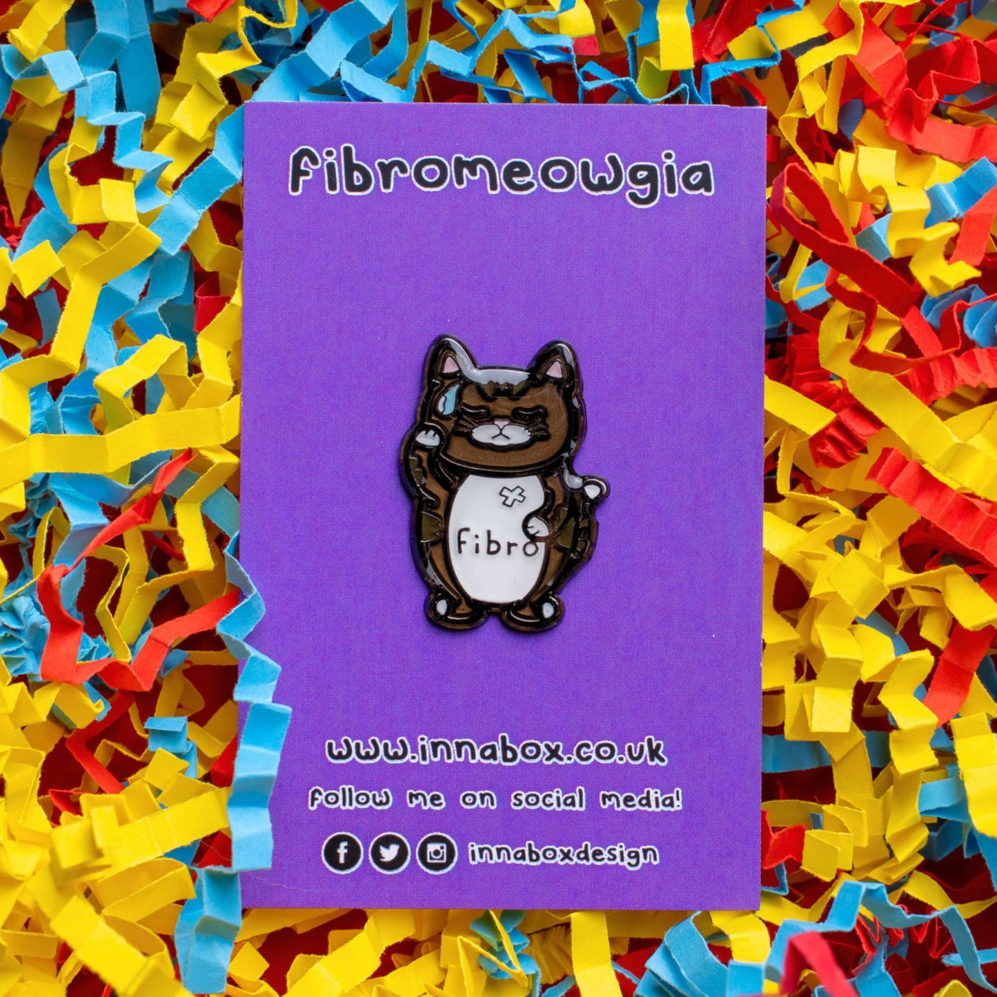 Fibromeowgia Enamel Pin - Fibromyalgia on purple backing card laid on a red, yellow and blue card confetti background. The enamel pin is a brown cat with a white stomach with fibro written across it. The cat has its eyes closed, a sweat droplet on its forehead and holding its head in its paw. The enamel pin is designed to raise awareness for fibromyalgia.