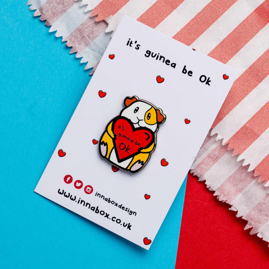 Guinea Pig Mental Health Enamel Pin on white backing card with red hearts laid on a blue and red card background. The enamel pin is of a smiling guinea pig holding a big red heart with the text it's guinea be ok inside. The enamel pin is deigned to raise awareness for mental health.